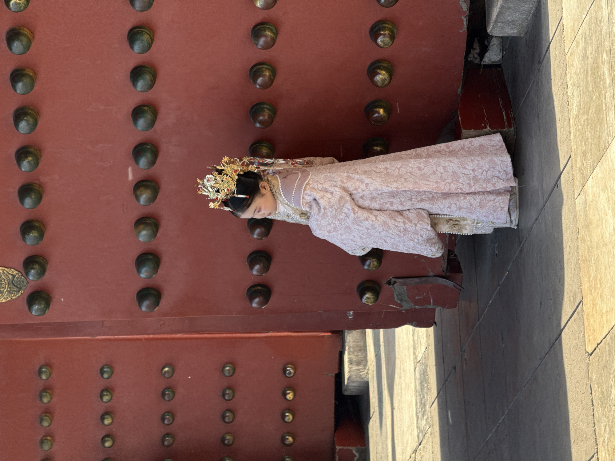 A woman in traditional Chinese clothing is standing next to a large red door adorned with metal studs. The door and its surrounding frame are situated next to a stone walkway. This scene is located in Dongcheng District, China.