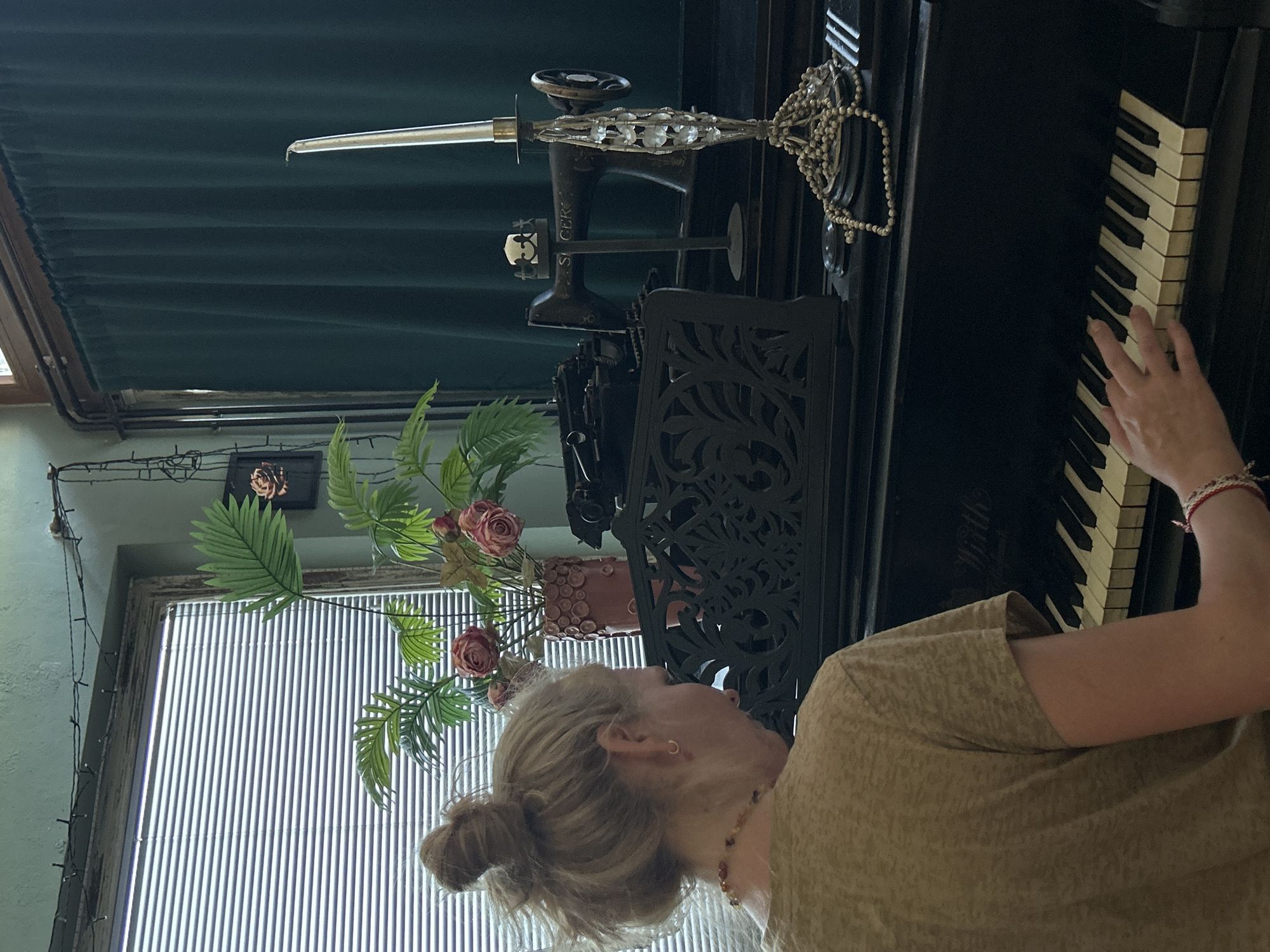 A woman with blonde hair pulled up in a bun is seen from behind, playing a black piano. She is wearing a tan t-shirt and a beaded necklace. Her right hand is on the piano keys. The piano has ivory and black keys. In front of the piano, there is an ornate black music stand. Behind the music stand, on the piano, sits an antique black sewing machine. To the right of the sewing machine, a tall, ornate silver candelabra holds a white candle. Draped around the candelabra and on the piano are strands of small, light-colored beads. Behind the piano, a dark green curtain is visible. To the left of the sewing machine and in front of the window, there is a vase with artificial pink roses and green foliage. The window behind the plants has horizontal blinds. The room has a greenish-blue wall and a wooden framed ceiling light fixture with some string lights draped across it.