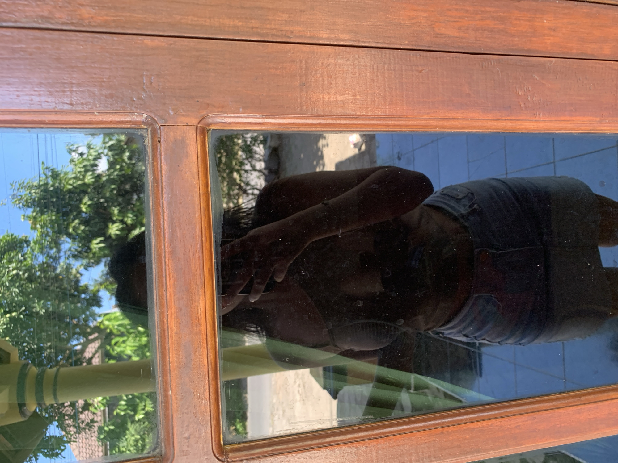 A full-body reflection of a person wearing a light-colored top and denim shorts is visible in a window pane. The person is holding a phone up to take the photo. The background reflected in the window includes trees, a building with a tiled roof, and a clear blue sky. The window is framed by a wooden structure with a reddish-brown stain. The foreground shows the wooden frame of the window. The image appears to be taken outdoors, possibly in a residential area.