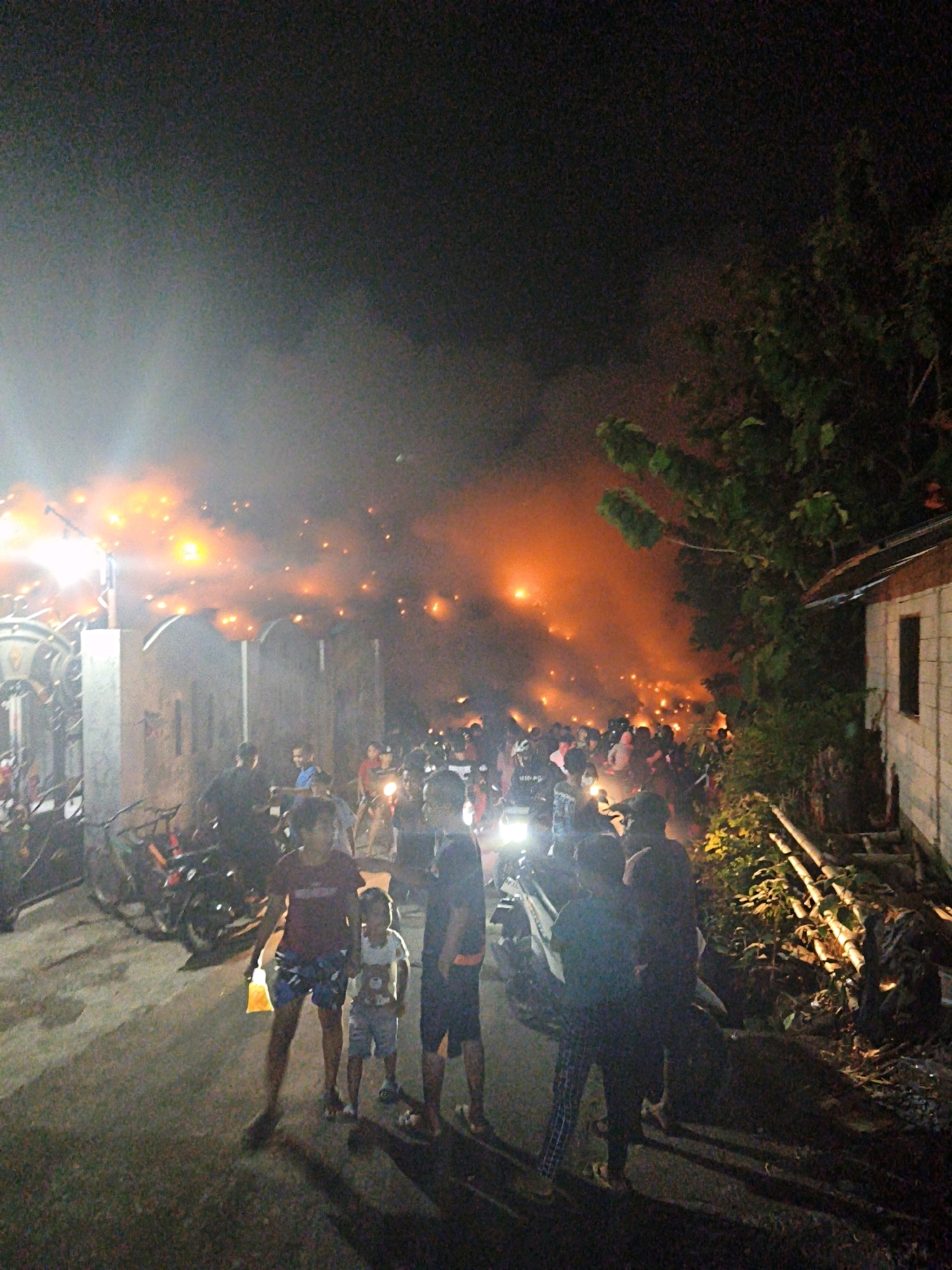 The image depicts a nighttime scene in the city of Surakarta, Indonesia, characterized by a large fire with orange flames and smoke engulfing the background. A crowd of people is gathered, some observing the fire, while others are standing or moving around. Several motorcycles are visible in the foreground and middle ground, some parked and others appearing to be in use. A building with a white wall and a dark window is present on the right side of the image, and trees with foliage are also visible. The ground is paved, and there are shadows cast by the artificial lights and the fire. A young boy in the foreground is holding a yellow object. The overall atmosphere is chaotic, with the bright flames and smoke suggesting a significant event.