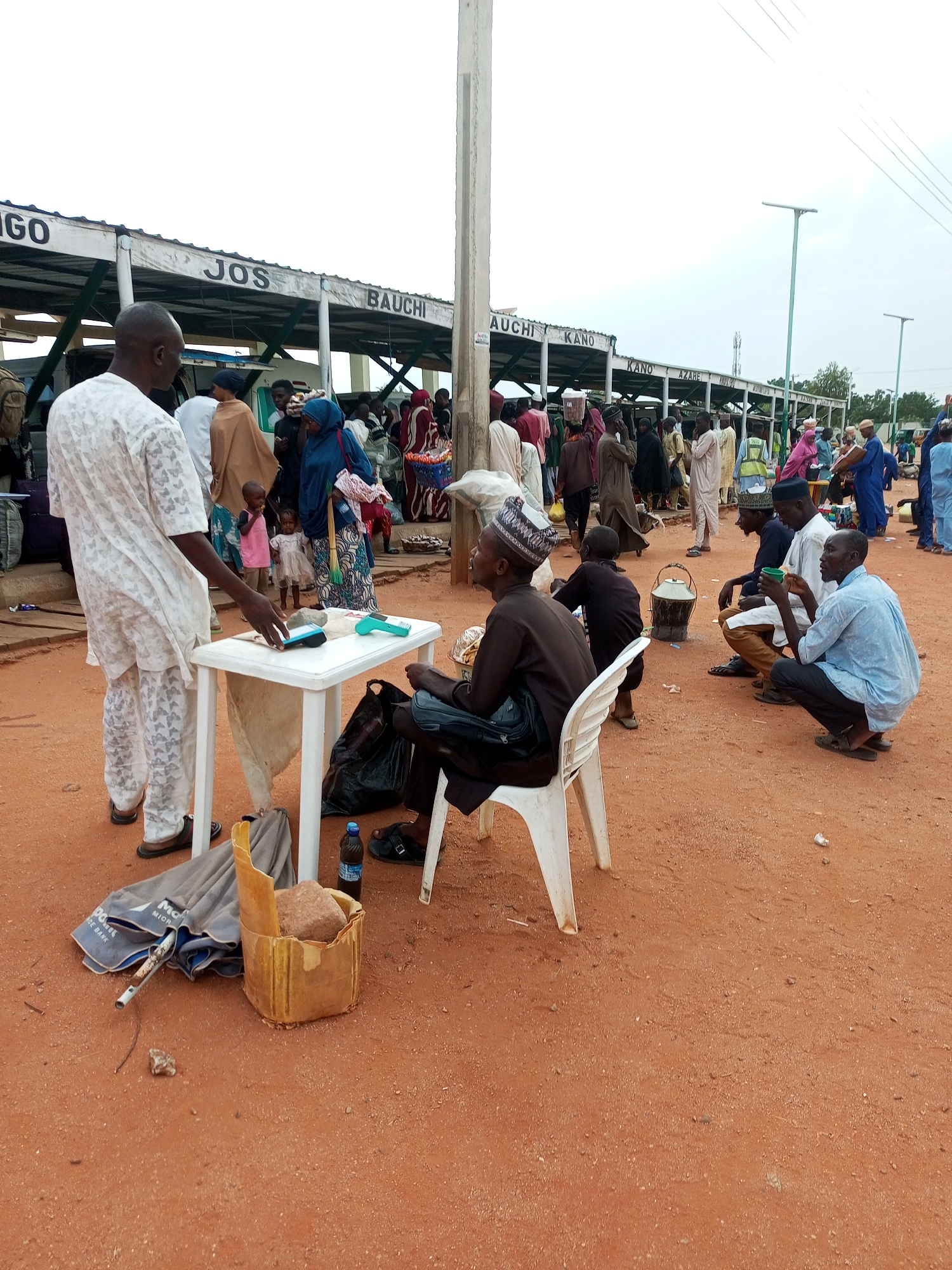 The image depicts a busy outdoor scene at a transportation hub, likely a motor park in Gombe, Nigeria. In the foreground, a man in a white patterned traditional outfit stands near a white plastic table. On the table are a transaction device, a stone, and a small blue and white machine. To the right, a man in a brown outfit sits on a white plastic chair, holding a cup and observing the scene. Another man in a light blue shirt squats beside him, also holding a cup.
The background reveals a covered waiting area with signs indicating various destinations, including "INGO", "JOS", "BAUCHI", and "KANO". Numerous people, some carrying luggage, are gathered under this structure and in the open area, suggesting a point of departure or arrival for travelers. The ground is covered in reddish-brown soil, typical of many parts of Nigeria. The overall atmosphere appears to be one of lively activity and waiting. A partially unfurled umbrella and a woven basket containing a stone are visible on the ground near the table.