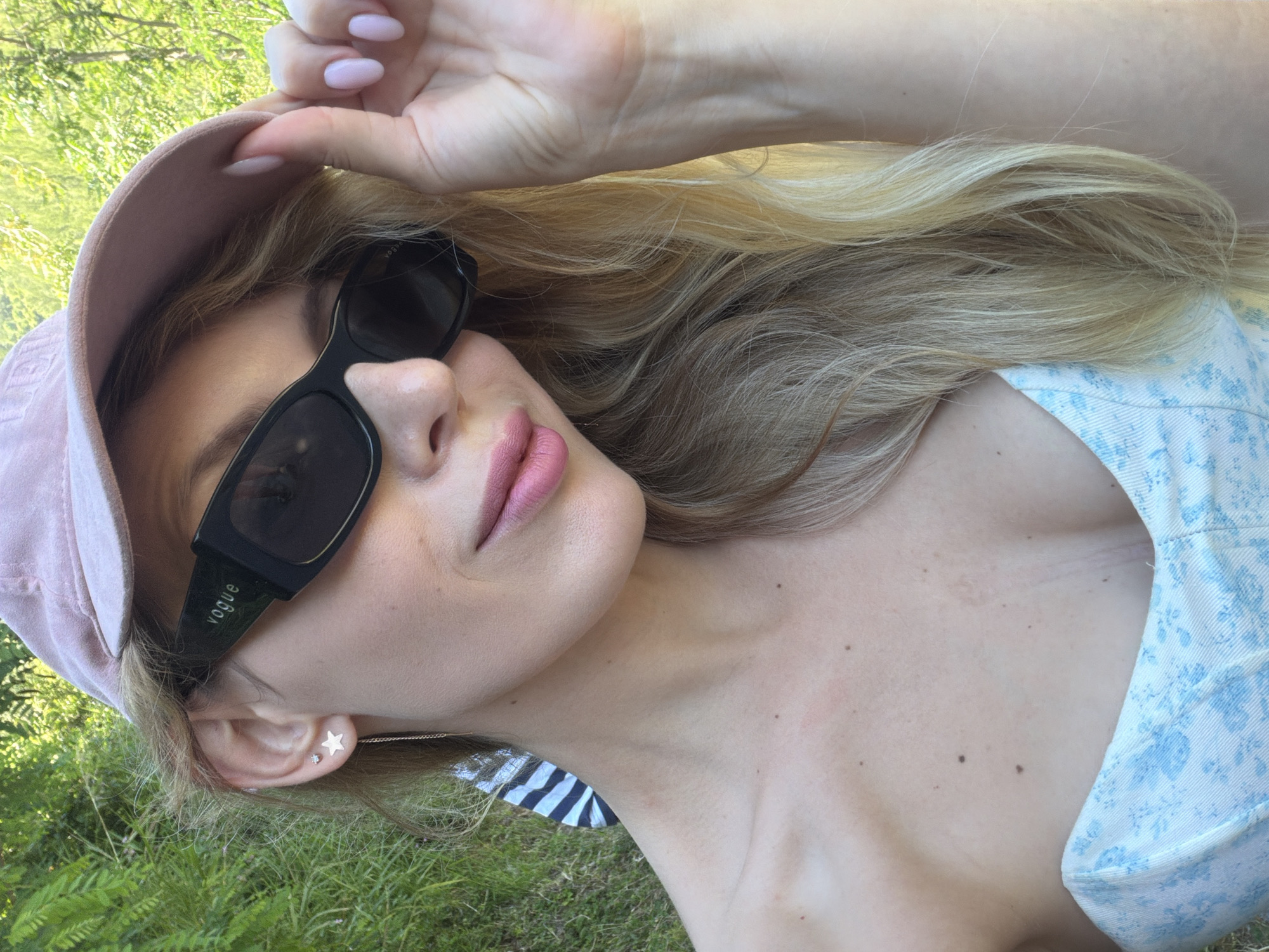 A fair-skinned female is depicted in a close-up, chest-up shot, outdoors in Budva, Montenegro. She wears a light pink baseball cap and black rectangular sunglasses with the word "vogue" visible on the left temple. Her long, wavy blonde hair is partially visible. Her left ear features two earrings: a small, clear stud in the upper lobe and a larger star-shaped stud with a slender chain dangling from the lower lobe. Her lips are tinted with a light pink color. Her right hand, with light pink polished fingernails, lightly touches the brim of her cap on the right side of her head. She wears a light-colored, possibly strapless, top with a blue floral pattern. The background consists of dense, out-of-focus green foliage, illuminated by bright daylight.