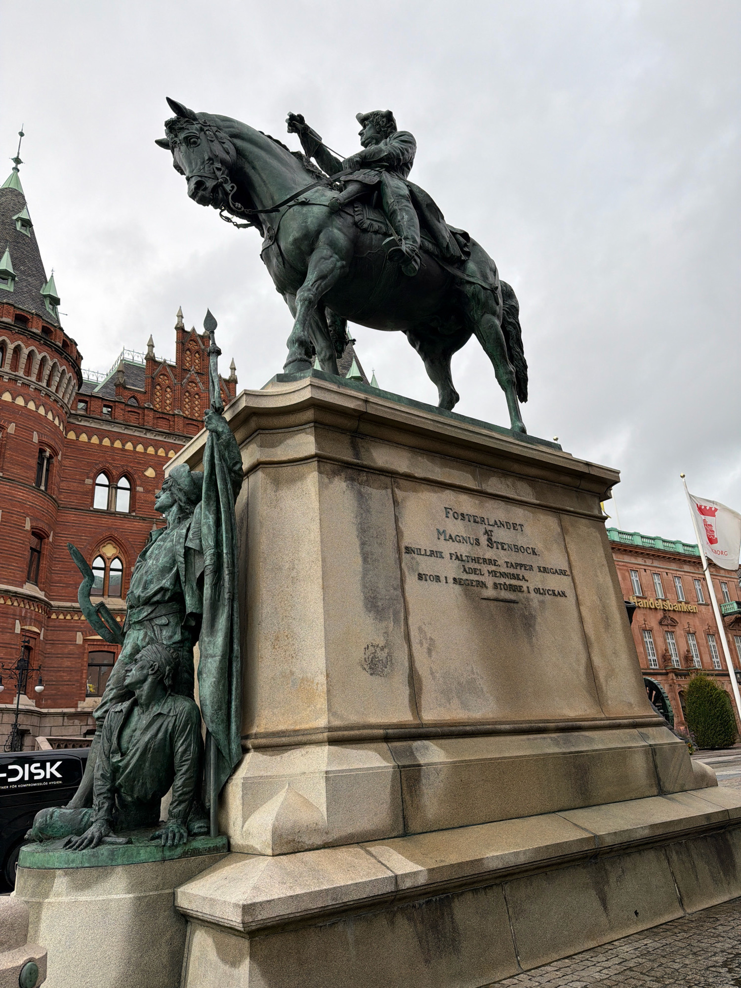 The image shows an outdoor, low-angle shot of a monumental equestrian statue and its base, located in Helsingborg, Sweden. The statue is made of bronze and depicts a man in military attire on horseback, holding a sword. The base of the statue is made of granite and features inscriptions, as well as smaller bronze figures at its base, including a kneeling man holding a banner and a spear.
To the left of the statue, a multi-story red brick building with ornate architecture and a pointed, green-roofed tower is visible. To the right, another building, also with red brick and white window frames, appears to be a commercial establishment, indicated by a sign that reads "Handelsbanken". A white flag with a red emblem is also visible to the right. The ground in the foreground is paved with cobblestones. The sky is overcast and gray.
The inscriptions on the base of the statue are in Swedish and honor Magnus Stenbock. The text reads: "FOSTERLANDET AT MAGNUS STENBOCK, SNILLRIK FÄLTHERRE, TAPPER KRIGARE, ÄDEL MENNISKA. STOR I SEGERN, STÖRRE I OLYCKAN." This translates to: "THE FATHERLAND TO MAGNUS STENBOCK, CLEVER GENERAL, BRAVE WARRIOR, NOBLE PERSON. GREAT IN VICTORY, GREATER IN MISFORTUNE." The statue is a prominent landmark and is situated in a public square or plaza.
