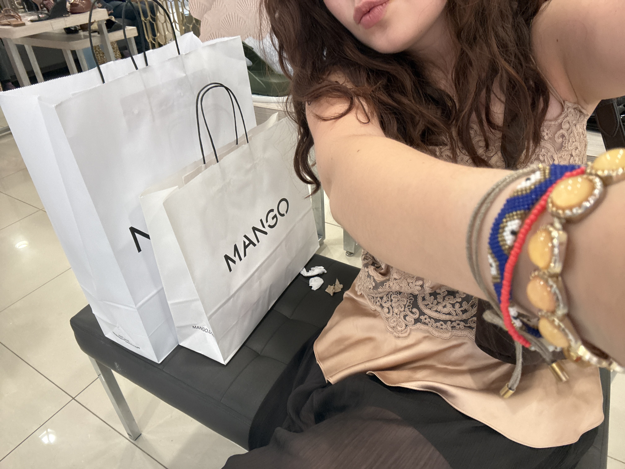 A medium indoor shot depicts a person posing for a selfie in Mexico City, Mexico, showcasing shopping bags.
The person is seen from the chest up, with dark brown curly hair and pink lips. She wears a beige lace-trimmed top, and her arm is adorned with several bracelets.
Two shopping bags are visible next to the person on a black seat. One bag has "Mango" printed on it, indicating a purchase from the Mango store. The second bag has only a "V" on the bag. A few scraps of paper are on the seat next to her.
The background includes a glimpse of other shoppers and retail displays, suggesting the photo was taken inside a shopping mall or department store. The environment suggests a bustling commercial area, which aligns with Mexico City's status as a major urban center.