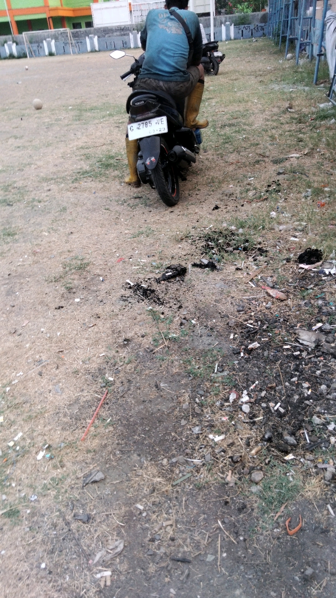 The image depicts a street scene in Tegal, Indonesia. In the foreground, a person is seated on a black Yamaha motorcycle, facing away from the camera. The rider is wearing a blue long-sleeved shirt, dark shorts, and yellow boots that extend up to their knees. The motorcycle has a license plate with the characters "G 2785 VE" and a date "03-29". The environment is a dry, dusty area with sparse vegetation. Scattered across the ground are cigarette butts, ash, and debris, suggesting a somewhat unkempt or neglected public space. In the background, a sports field with a goalpost and a white ball can be seen, along with some buildings that appear to be part of a recreational facility. A blue metal fence is also visible on the right side of the image, bordering the area. The lighting suggests it is daytime, with the sun casting shadows.