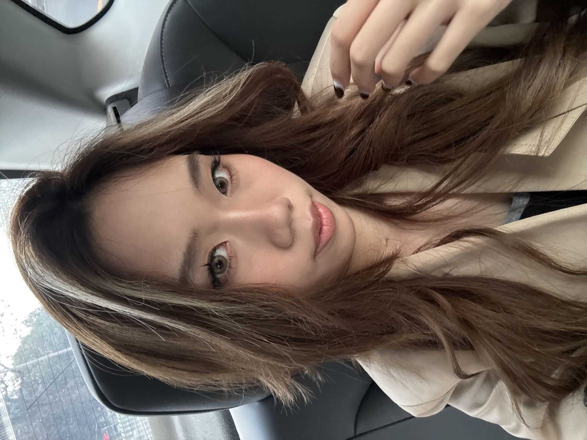 A young Asian woman is photographed from a high-angle perspective, seemingly within a car. She has long, wavy brown hair with blonde highlights. The woman's hand is visible near her hair, with painted nails. She has light skin, brown eyes, and pink lips. She is wearing a light beige jacket or coat. The background includes the car's interior with a black leather seat and a glimpse of the car window and rear-view mirror. There is no evidence to indicate the location is Lanzhou, China.