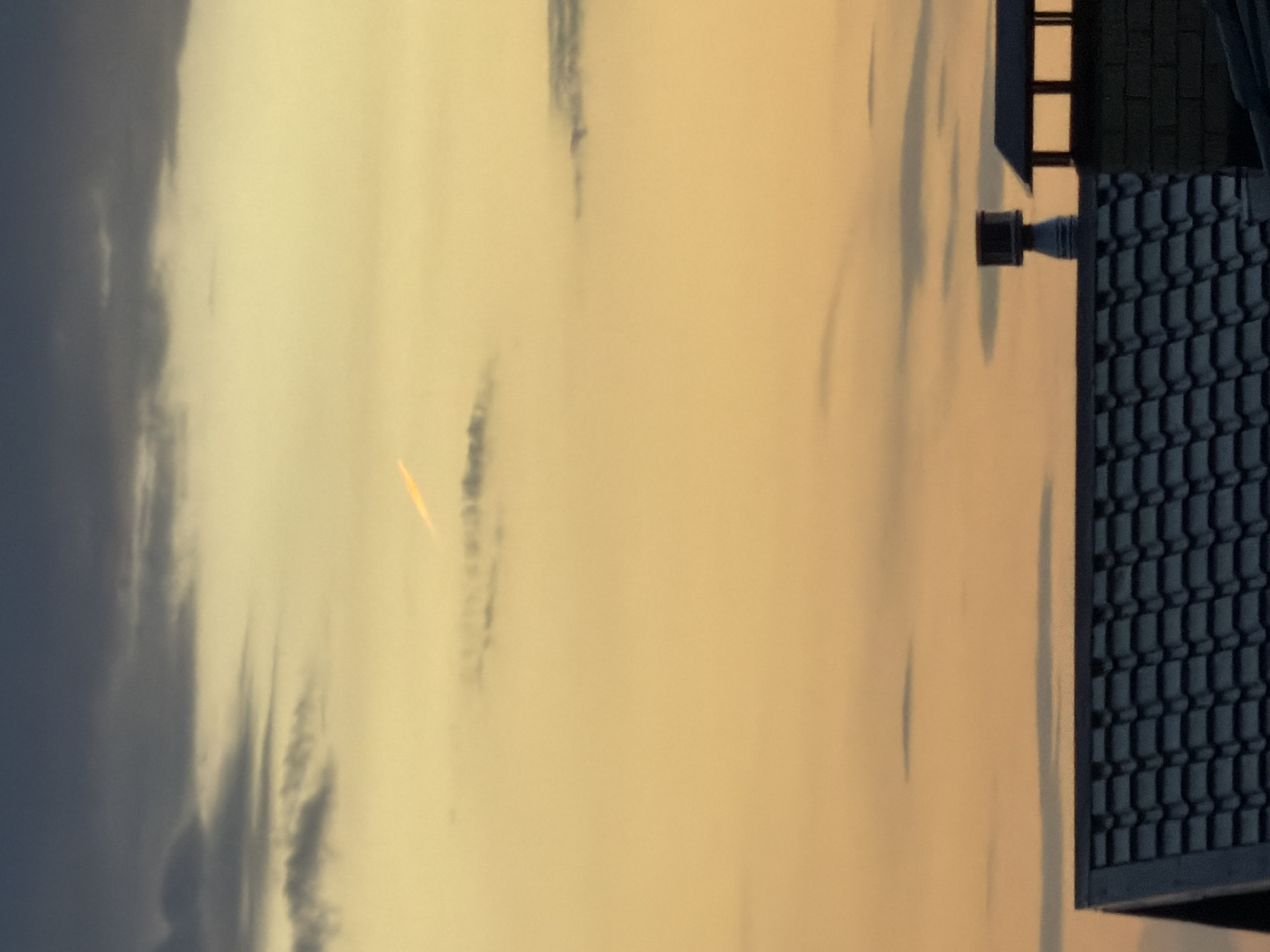 The image shows a view of the sky during what appears to be sunrise or sunset. The sky has a golden-yellow hue, with some dark clouds visible on the left side and scattered throughout. On the right side of the image, a part of a building is visible, featuring a dark blue tiled roof, a chimney, and part of a grid-like structure on the side. The location is possibly the city of Chaikovsky, Russia, but there is no explicit evidence of it in the image.