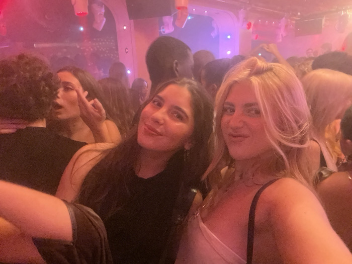 The image depicts a crowded indoor nightlife establishment, likely a nightclub or bar, situated in Paris, France. The scene is illuminated by artificial pink and red lighting, creating a hazy atmosphere.
In the foreground, two young adult females are prominent. On the right, a blonde-haired female with wavy hair looks directly at the camera, her lips puckered. She wears a light-colored, possibly white, strapless top with a thin dark strap visible over her right shoulder and multiple necklaces. To her left, a dark-haired female with long, straight hair also looks at the camera with a slight smile, wearing a dark-colored top.
Behind these two and slightly to the left, another dark-haired female is visible, positioned in profile facing away from the camera. She is seen with her mouth open and her right hand raised, extending her middle finger upwards. A person with dark, curly hair, their back to the camera, is partially visible in the far left foreground.
The background is densely populated with numerous other individuals, largely out of focus, suggesting a large gathering. Overhead, the ceiling displays various light fixtures and decorative elements, some appearing as suspended white objects. A curved architectural feature with internal lighting is visible towards the left background, framed by the pink and red illumination.