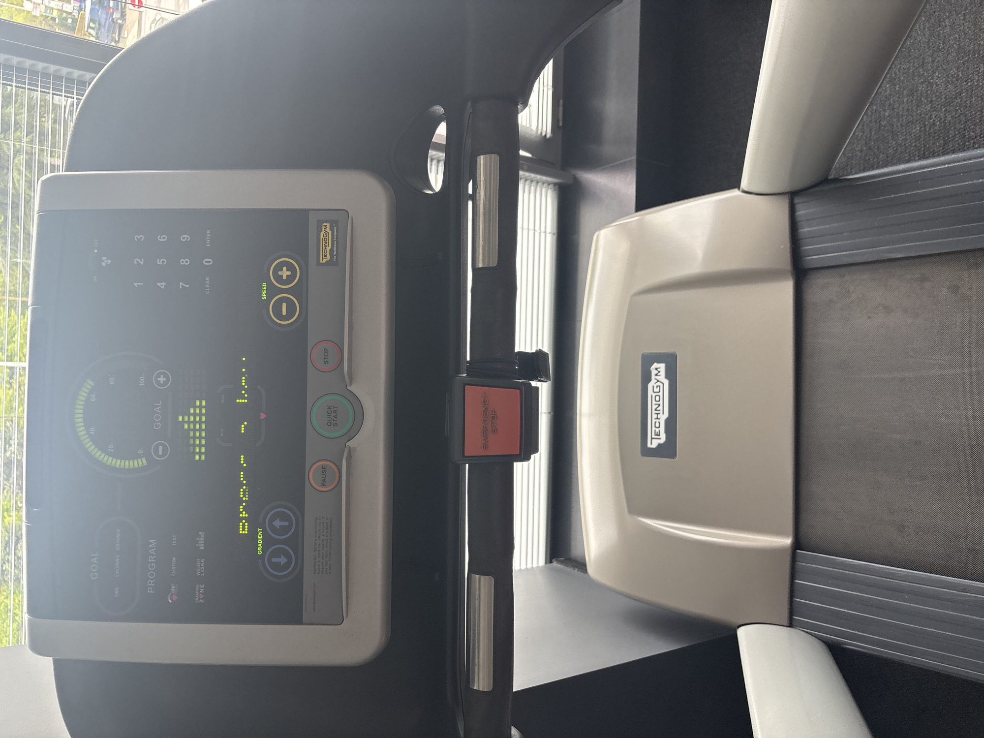 The image displays a modern Technogym treadmill in what appears to be a gym setting in Blagoevgrad, Bulgaria. The treadmill is viewed from above, showing its control panel, handrails, and part of the running belt. Natural light streams in from a window visible on the left side, suggesting it's daytime.
The control panel features a digital display with various workout options and metrics. Visible text on the screen includes "GOAL" (with options like CALORIES and DISTANCE), a circular progress bar, and bar graphs. Below this, "PROGRAM" is displayed, offering choices such as "CRM," "CUSTOM," "TEST," and "WEIGHT LOSS." Physical buttons include "SPEED" with '+' and '-' controls, and "GRADIENT" with up and down arrows. A numeric keypad (0-9 with CLEAR and ENTER) is also present. To the right of the screen are "QUICK START," "PAUSE," and "STOP" buttons. A prominent red "EMERGENCY STOP" button is centrally located on the handlebar. The brand "TECHNOGYM The Wellness Company" is clearly visible on the screen and the main body of the treadmill.
No people are visible, and the treadmill appears to be idle or paused, ready for use. A partial warning message is visible: "WARNING Before exercising check up to."