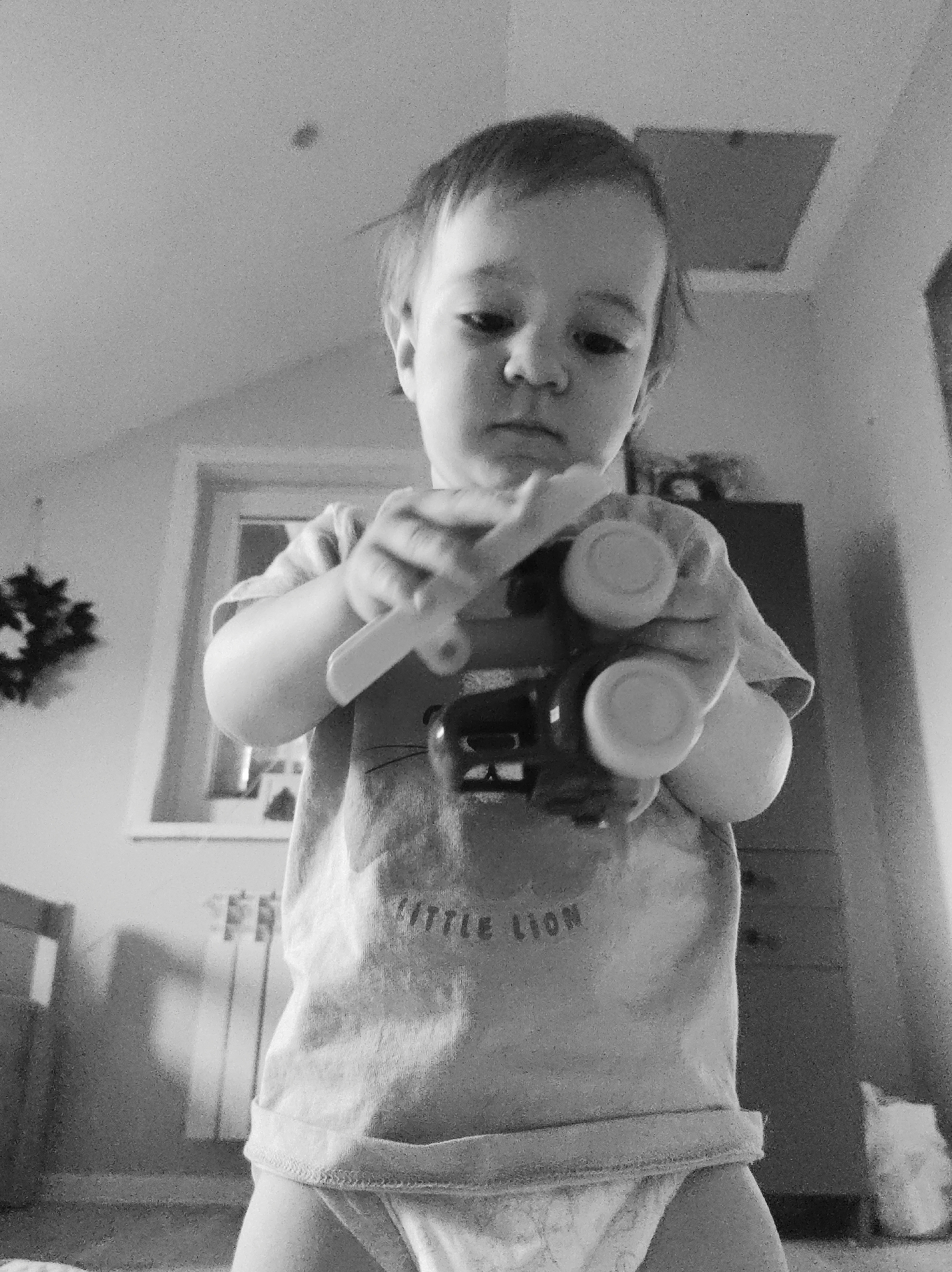 A young child stands indoors, holding a toy vehicle with large wheels. The child wears a t-shirt with the words "LITTLE LION". A window with a wreath, a radiator, and a cabinet are visible in the background. The room appears to be in a residential location, city and state Unknown.