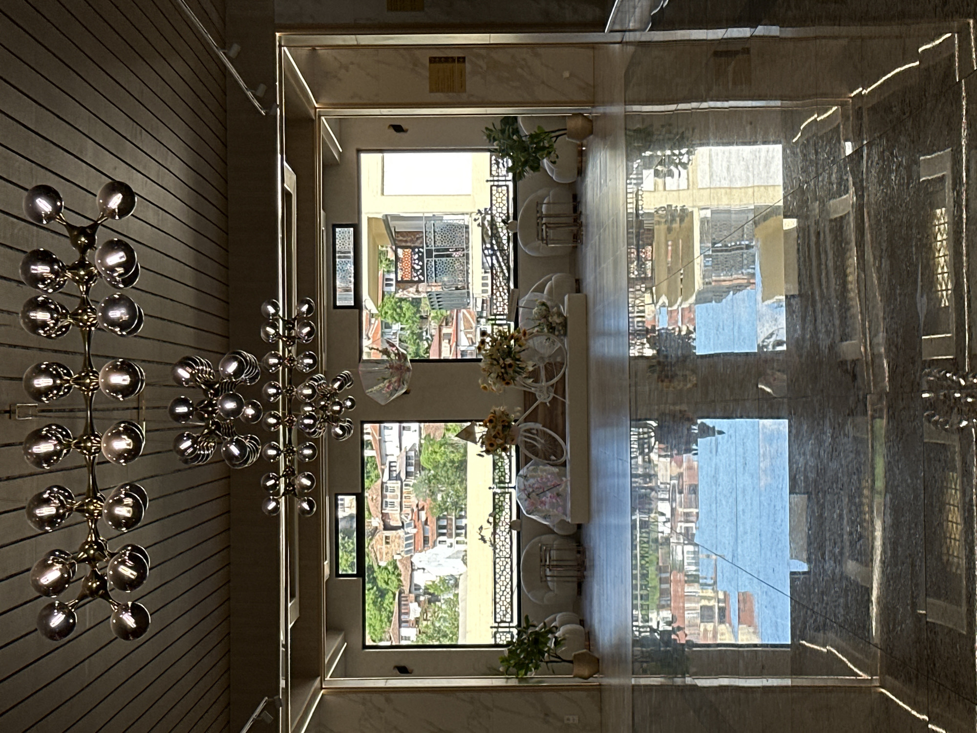 The image displays the interior of a spacious room with a panoramic view of the city of Veliko Tarnovo, Bulgaria, through a large rectangular window.
The ceiling features light-colored, horizontal wooden slats from which multiple contemporary chandeliers are suspended. The prominent chandeliers are designed with metallic, spherical bulbs arranged in a branching, molecular structure. The floor is a highly polished, dark, reflective surface, mirroring the interior elements and the exterior landscape with notable clarity.
In the midground, directly in front of the large window, a seating arrangement and decorative elements are present. Two rounded, plush, off-white armchairs flank a pair of nested, metallic (gold-toned) round side tables. Two white decorative bicycles, adorned with floral arrangements, are positioned on either side of the central axis; one features sunflowers, and the other is partially covered with a sheer, floral-patterned fabric. Two potted green plants in gold-toned planters are situated on the floor to the left and right of this arrangement. A sheer decorative umbrella with floral patterns hangs from the ceiling in the upper right portion of the window area. The window is framed by light-colored, possibly marble or stone, panels accented with gold trim.
Through the large window, a dense collection of traditional houses is visible, characterized by white or light-colored facades and red-tiled roofs, built into a green hillside. Green trees and foliage are interspersed among the structures. A body of water or light-colored ground area is visible in the middle distance, separating the interior from the distant cityscape. A decorative dark metal railing with geometric patterns is positioned directly in front of the window at waist height. No individuals are present in the scene.