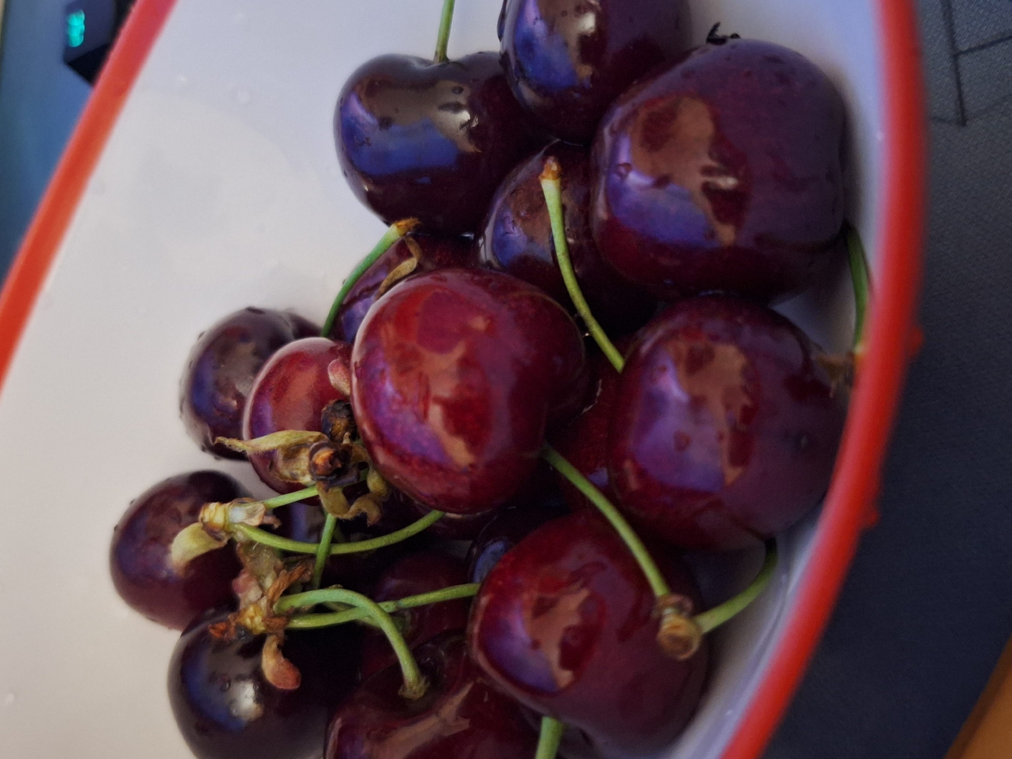 The image shows a close-up, high-angle shot of a bowl of dark red cherries. The cherries are plump and glossy, with some appearing to have water droplets on their surface. Green stems are visible, attached to several of the cherries, with some dried, brown remnants of where the cherries were previously connected to the branch. The bowl is white with a red rim and is placed on a dark surface, possibly a table or countertop. The background is out of focus, but a hint of a blue and green electronic device is visible in the upper right corner. The image quality is sharp, capturing the texture and sheen of the fruit. There are no people or animals present in the image. This image was taken indoors, likely in a residential kitchen or dining area.