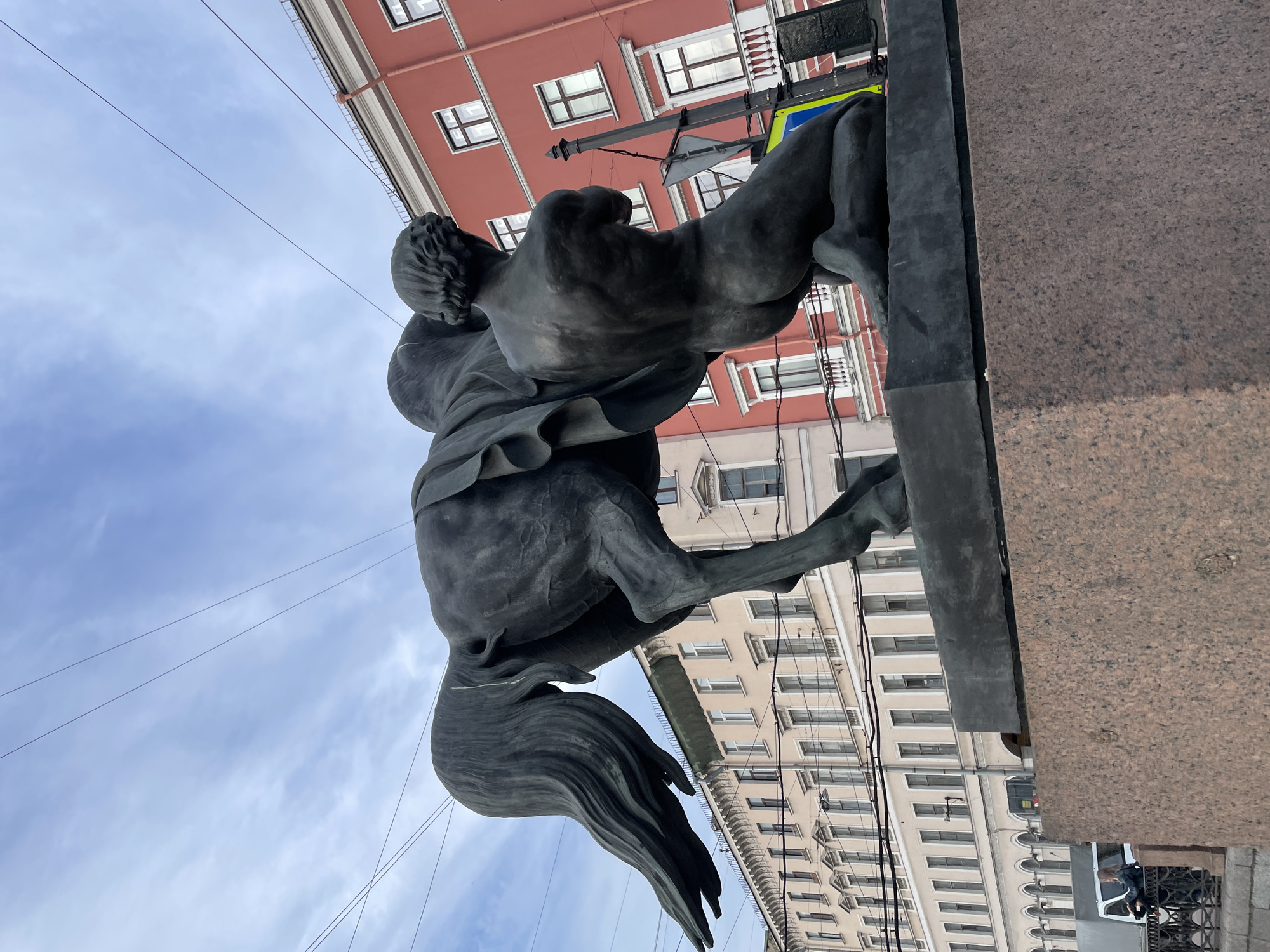 A low-angle shot captures a dramatic equestrian statue against an urban sky and buildings in Saint Petersburg, Russia. The statue, made of dark, weathered metal, depicts a powerful horse rearing dynamically on its hind legs. Its rider, a male figure, is poised, looking slightly upward and to the side, conveying a sense of intensity or contemplation. The horse's tail is long and flowing, adding to the sense of movement.
The statue is set upon a large, textured pedestal of reddish-brown granite, which occupies the lower right portion of the frame. In the background, under an overcast sky, are multi-story buildings. One prominent building features a red facade with evenly spaced rectangular windows. Another building, partially visible behind the horse, is cream-colored with numerous windows and decorative cornices, hinting at classical architecture.
Several thin utility lines crisscross the overcast sky, visible above and between the statue and buildings. The lighting suggests daytime, but the clouds obscure direct sunlight. The dynamic pose of the horse and rider creates a sense of action and monumentality. No discernible text is visible on the statue or the surrounding structures.