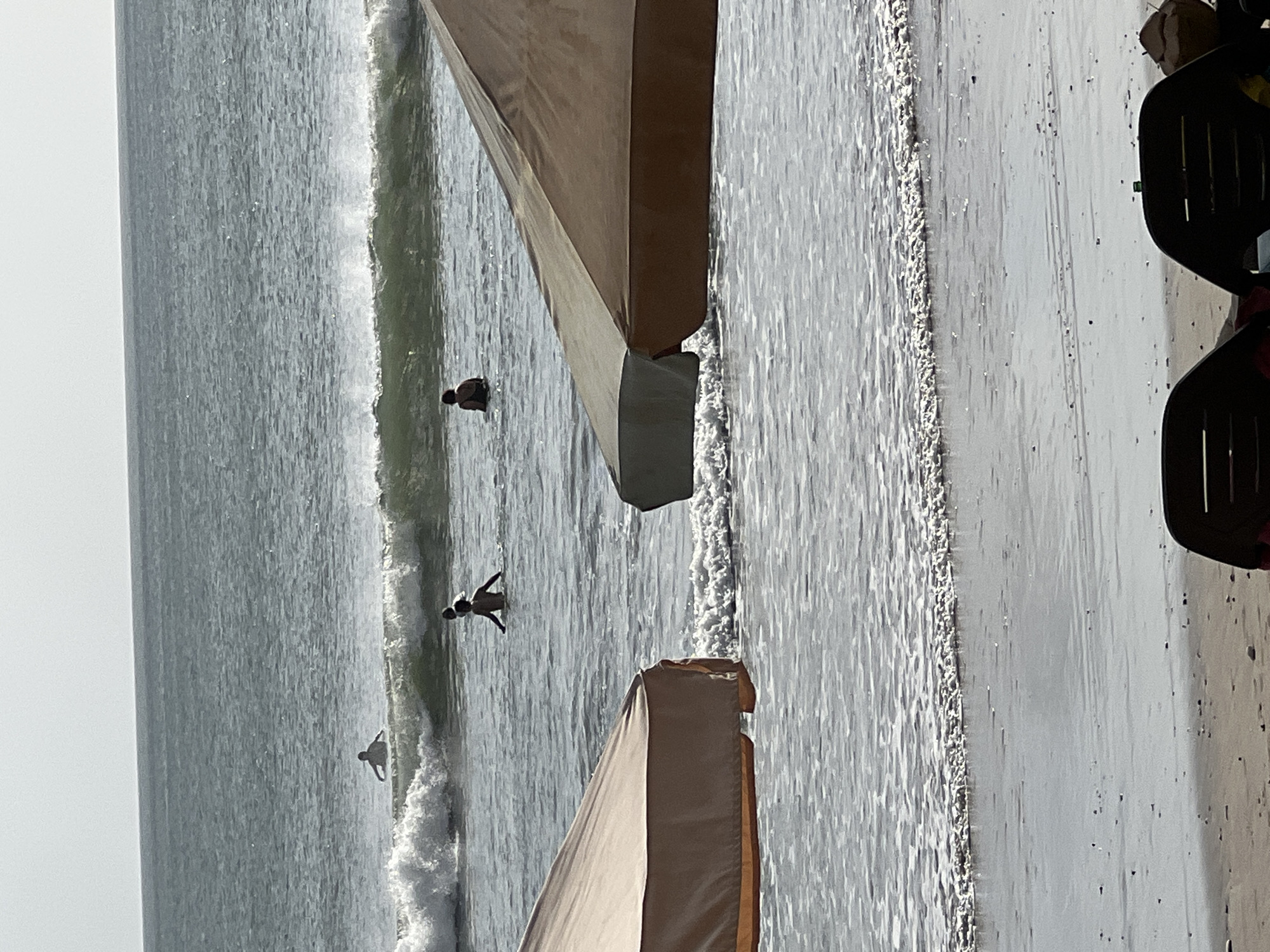 The image shows a beach scene in Kuta, Indonesia. In the foreground, two beach umbrellas are partially visible, with one being a tan and cream color and the other appearing darker. The umbrellas cast shadows on the wet sand. The ocean occupies the middle ground, with gentle waves breaking near the shore. Several people are in the water; one person is wading, and two others appear to be swimming or standing in the surf. A fourth person is further out in the water, possibly on a surfboard. The horizon is visible in the background, with a clear, bright sky above the ocean. The overall impression is a relaxed beach day.