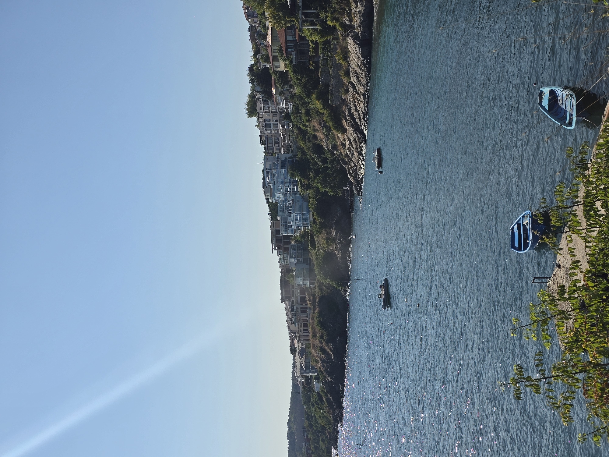 This is a scenic view of the sea in Sozopol, Bulgaria, with the coastline lined with buildings. The water is blue and calm, reflecting the sunlight. In the foreground, two small blue rowboats are tied to a concrete pier, partially obscured by green foliage. Further out in the water, a larger gray boat is visible. The coastline is rocky and covered with green vegetation and trees. Several multi-story buildings, some appearing to be under construction, are situated on the hillside overlooking the sea. The sky is clear and blue. A diagonal light streak is visible in the upper left portion of the sky, likely caused by the sun's rays.