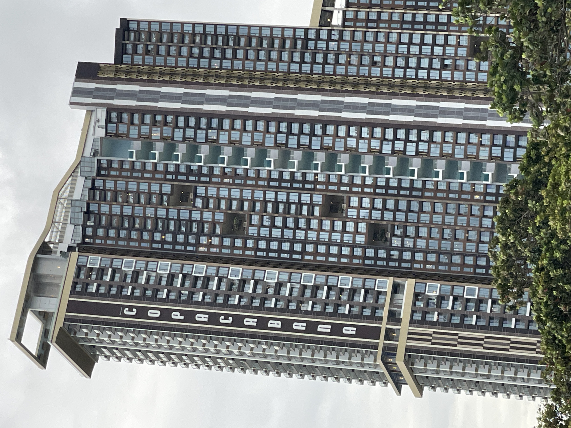 The image shows a tall, modern condominium building in Pattaya City, Thailand, with the word "COPACABANA" written vertically on its facade. The building features a dark brown and white color scheme, with many windows and balconies. Green trees are visible in the foreground. The sky is overcast.