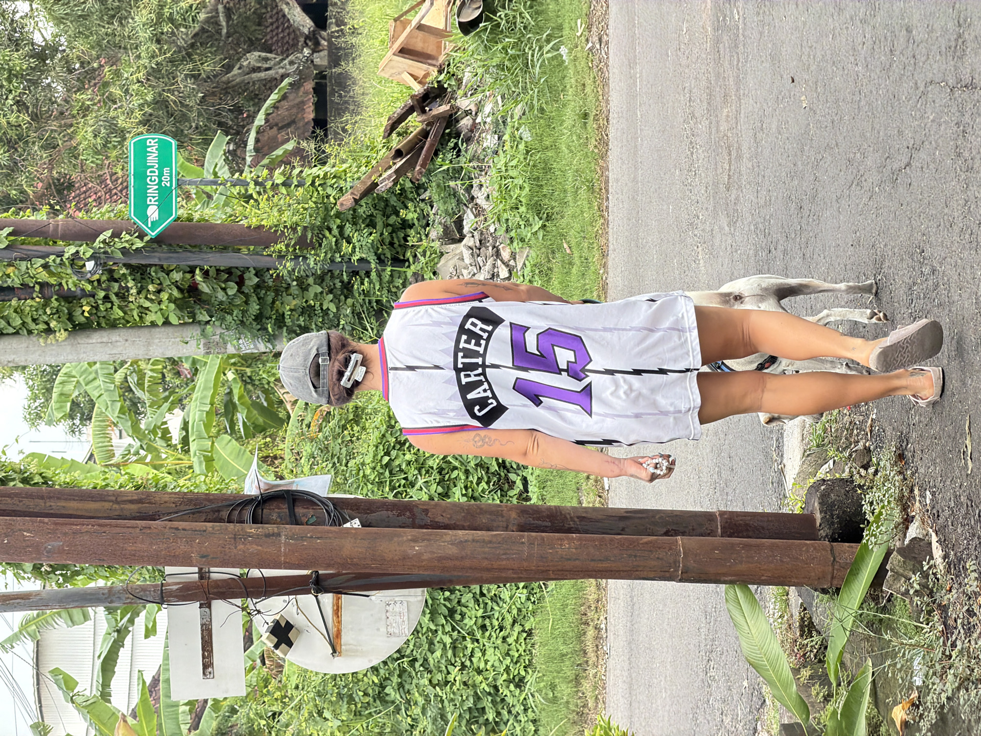 In North Kuta, Indonesia, a woman wearing a white basketball jersey with "CARTER" and the number "15" on the back, along with a grey baseball cap, walks down a paved street. She is holding a white beaded bracelet in her right hand. A white dog with a blue collar is walking beside her. The street is lined with lush green vegetation, including large banana leaves and vines. A rusty metal pole with wires attached stands on the left side of the road, and a green directional sign pointing to "RINGDJINAR 20m" is visible on the right. Debris, including broken wood and rocks, is scattered in the background. The sky is overcast, suggesting a cloudy day.