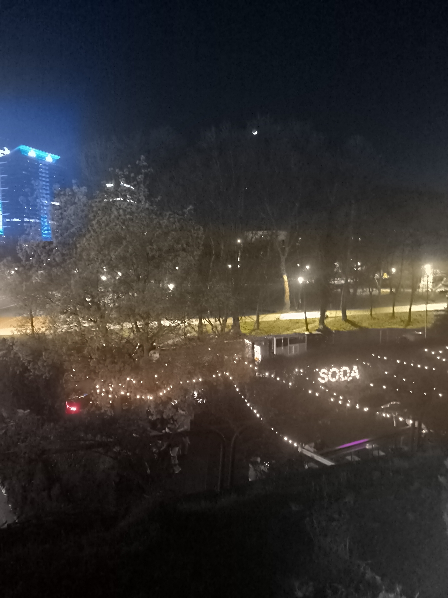 At night, a cityscape in Sofia, Bulgaria, features a tall, illuminated building on the left. A crescent moon is visible in the sky. A park-like area with trees is in the foreground, decorated with strings of lights. The word "SODA" is spelled out with lights. A road with car headlights can be seen.