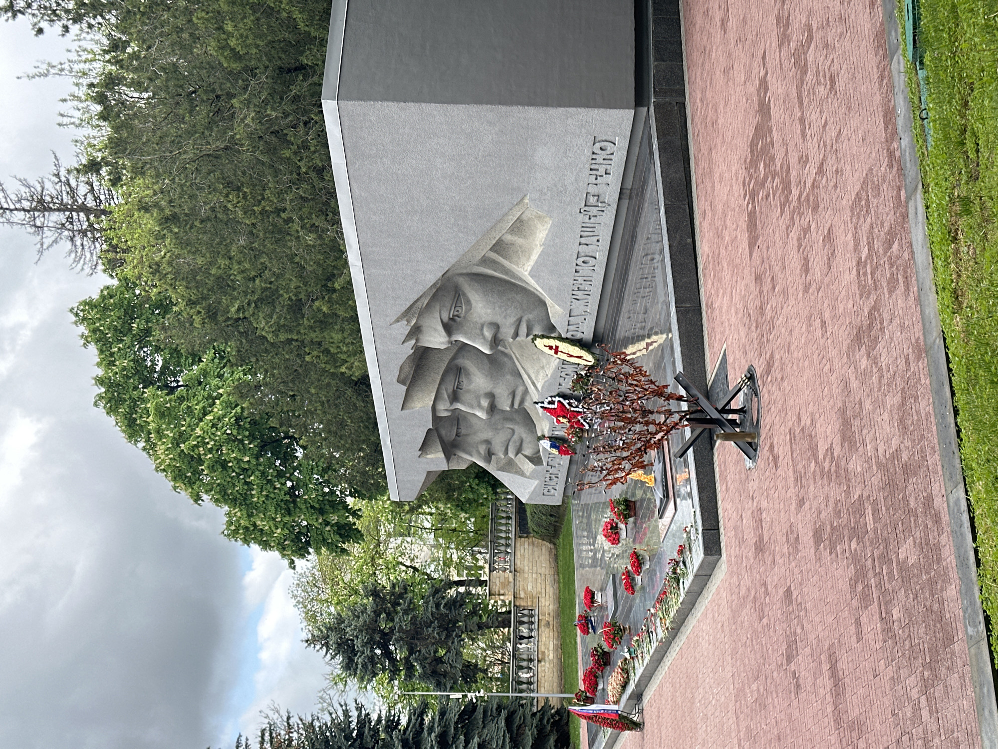 The image shows a memorial in Stavropol, Russia, dedicated to soldiers. The memorial features two carved heads in a gray stone structure with inscriptions in Russian. Red and white wreaths and flowers are placed at the base of the monument. A metal, star-shaped structure with dry branches stands among the flowers. The background includes trees and a brick-paved walkway. The sky is overcast.