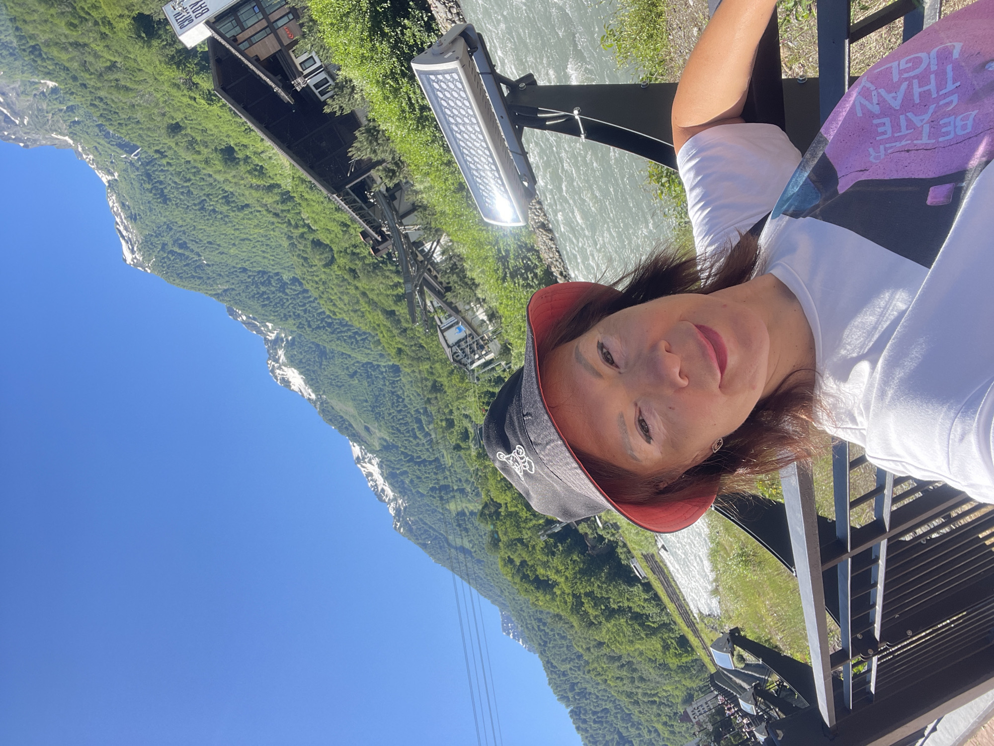 A woman is taking a selfie in a mountainous area with a river. She is wearing a white t-shirt, a black bucket hat with a red brim and a cartoon image on it, and pink lipstick. She has brown hair and is smiling. In the background, a river flows through a valley. Mountains covered in green trees and some snow are visible in the distance. There are buildings and a cable car system on the mountainside. A modern street light is mounted on a pole in the foreground. The scene appears to be in Estosadok, Russia, a resort town known for its mountain landscapes and ski infrastructure. The overall impression is a sunny day with a clear blue sky.