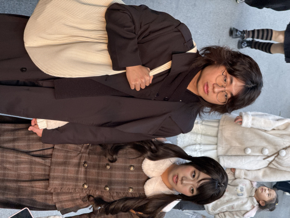 The image displays a street scene in Liangjiang, China, featuring multiple pedestrians in various states of movement or pause. In the immediate foreground, a male and a female individual stand side-by-side. The male, on the left, has dark, wavy shoulder-length hair and wears circular eyeglasses. He is dressed in a dark trench coat over a dark shirt and scarf, and carries a light-colored, vertically pleated tote bag over his right shoulder. His expression is neutral, and he gazes slightly to the viewer's left. The female, on the right, has dark hair styled into two pigtails. She wears a brown plaid dress or top with a white fur-trimmed peter pan collar and appears to be speaking or exhaling, with her mouth slightly open, looking slightly to the viewer's right. Their right hands are in close proximity, but no direct physical interaction is explicitly visible.
In the mid-ground, other individuals are partially visible. To the far left, a person's lower legs and feet are seen, wearing dark shoes and horizontally striped brown and black knee-high socks or leg warmers. Behind the foreground subjects, a person wears a light-colored, possibly cream or white, faux fur coat and a long, white skirt with a visible lace trim. To the right of this person, a young child is present, wearing a light-colored hooded jacket with ear-like features on the hood. The child is looking upwards and slightly to the viewer's right. The ground beneath the individuals appears to be a light-colored, even paved surface. The ambient lighting suggests daylight conditions.