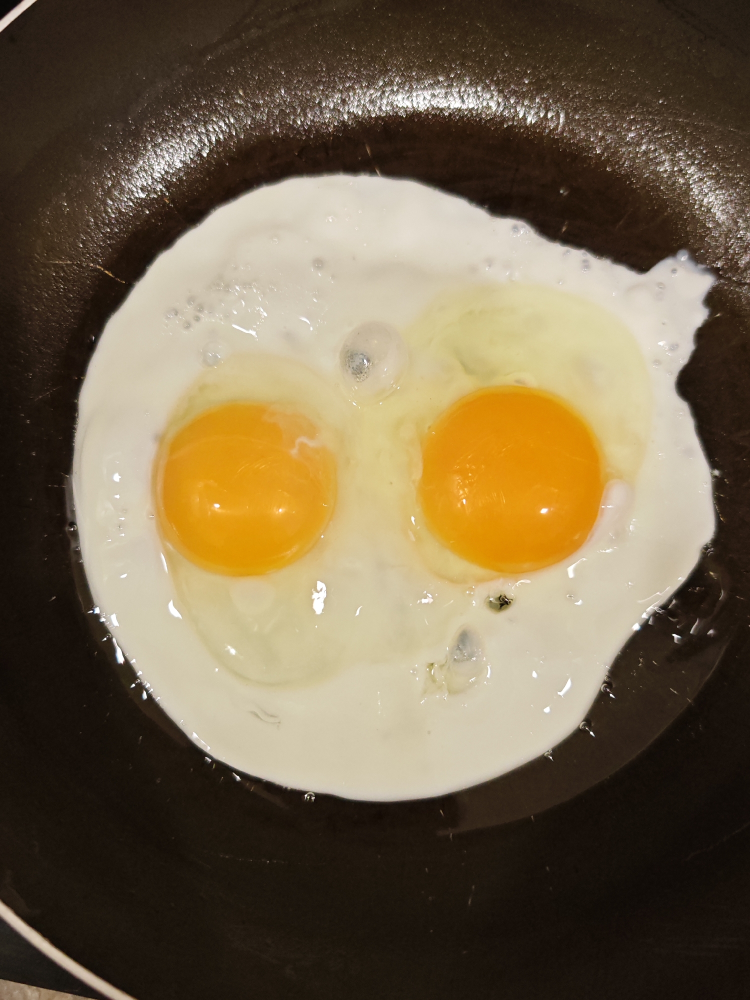 Two fried eggs are cooking in a dark-colored frying pan, likely non-stick. The eggs are sunny-side up, with intact, bright orange yolks and translucent white. Bubbles are visible in the egg whites, indicating they are actively cooking. The frying pan has a textured surface, likely the result of its non-stick coating. The overall lighting is overhead and somewhat diffused, creating highlights on the yolks and the surface of the pan. The scene is a close-up shot, focusing entirely on the food being prepared.
