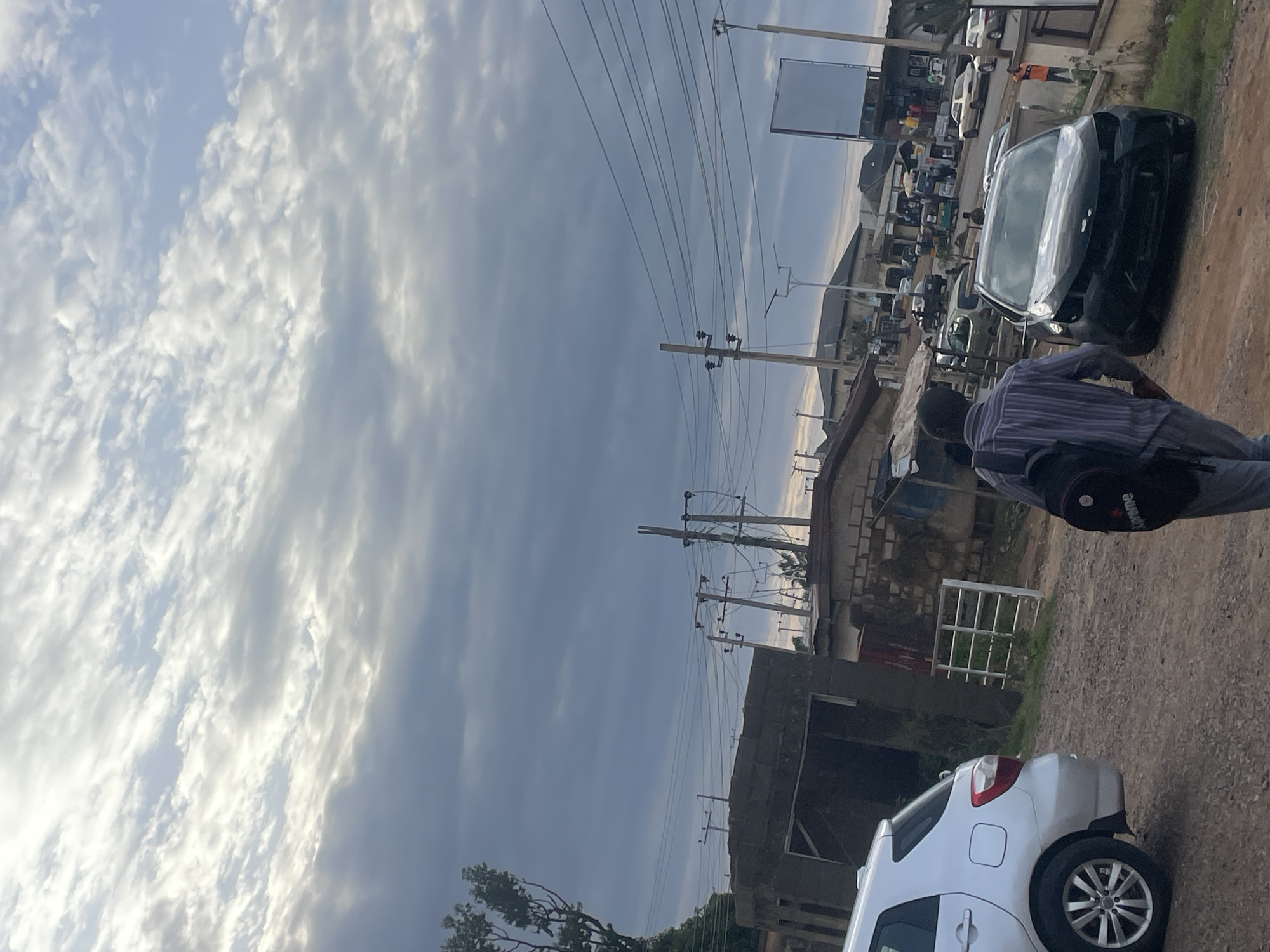 The image depicts a street scene in Rafin Par, Nigeria, under a cloudy sky. A man wearing a striped shirt and backpack is walking away from the camera towards the right of the frame. In the foreground, a silver car is parked on the left, and a damaged black car is positioned in the middle ground. The background shows several buildings, utility poles with electrical wires, and other vehicles on the road, suggesting a commercial or residential area. The overall impression is of a slightly overcast day in an urban environment.