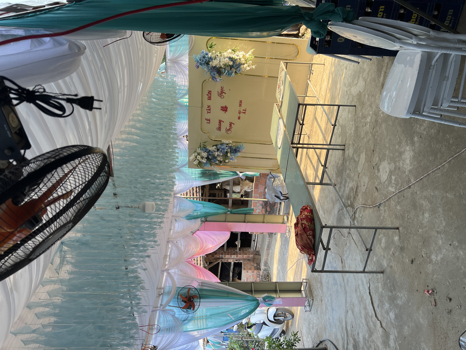 This image captures an outdoor or semi-outdoor event setup, likely in preparation for a celebration, in Bình An, Vietnam. The scene is adorned with festive white, light blue, and hints of pink fabric drapes creating an elaborate canopy overhead.
A prominent feature is a yellow-cream colored backdrop, decorated with elegant floral arrangements of blue and white flowers. Visible text on this backdrop reads "LÊ TÂN HÔN", which translates to "Engagement/Wedding Ceremony". Below this, the names "QUANG & NGỌC" are inscribed, followed by "TRỌNG". A specific date, "06 - 03 - 2026", is clearly displayed, along with "AL", likely indicating the Lunar calendar date.
In the foreground and extending towards the backdrop, several long, narrow tables with green tops and metal frames are arranged on a concrete floor. White plastic chairs are visible, some stacked and others positioned around the tables. A red patterned cloth is draped over one of the tables.
Multiple electric fans, featuring both black and orange blades, are strategically placed within the area, suggesting a need for ventilation. Power cords and plugs are visible, indicating the electrical setup for the event. In the background, partially constructed brick structures and a white scooter can be seen, providing context to the surrounding environment. The lighting, characterized by warm, directional sunlight casting long shadows across the concrete floor, suggests either late afternoon or early morning on a clear day. The overall impression is one of anticipation for a significant celebratory event. Partially visible text "on" and "eer" can be seen on stacked crates.