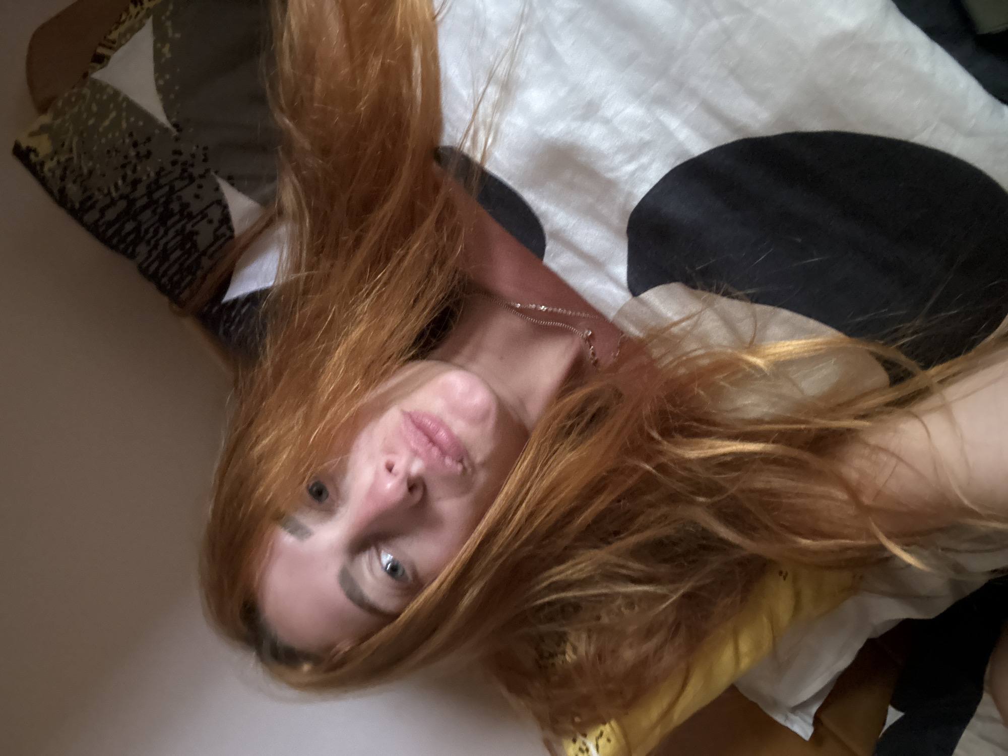 The image displays a close-up, angled portrait of a female subject in an indoor environment, located in Budva, Montenegro. The subject has long, wavy, reddish-auburn hair and fair skin. Her light-colored eyes are directed towards the camera. Her lips are slightly parted, with a noticeable texture on the upper lip. A reddish flush is visible on her nose and cheeks. She is wearing a delicate, thin, silver-toned chain necklace. The subject is positioned on a bed, with a yellow pillow supporting her head on the left side of the frame. She is partially covered by a white duvet or sheet patterned with large, irregular black ovoid shapes. Behind her head, a patterned pillow or cushion exhibits a geometric design comprising grey areas, black speckles, and white triangles. The background consists of a plain, light beige wall.