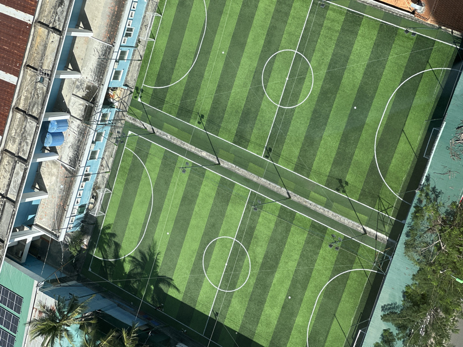 Aerial view of two adjacent artificial turf sports fields with dark and light green stripes. White lines define field boundaries, center circles, and penalty areas. A tall net and poles separate the two fields. Adjacent buildings flank the fields, including a blue and white structure and a green roofed building with solar panels. Trees are present on the lower left and right side of the image.