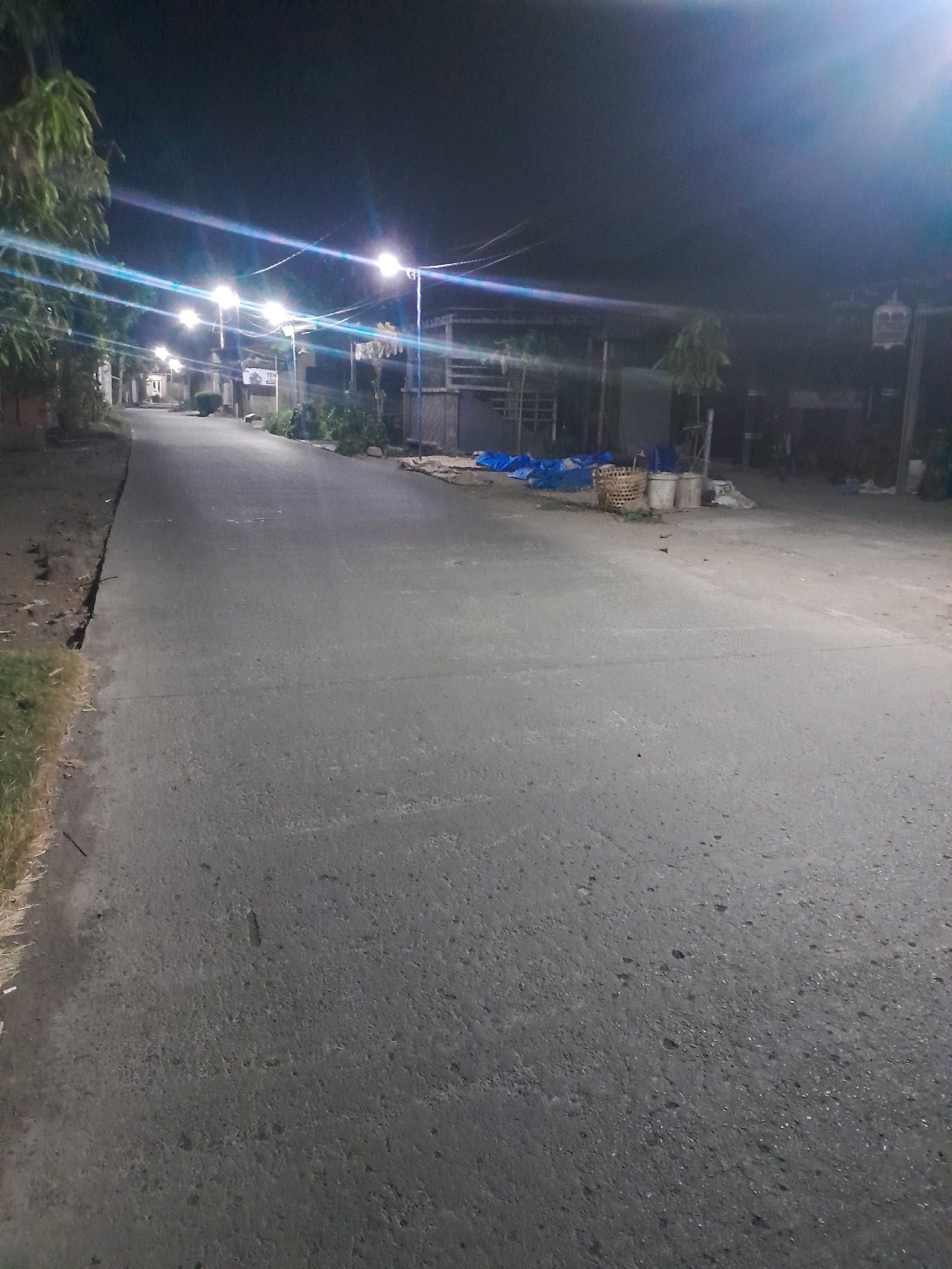 The image depicts a nighttime scene on a paved road in Tegalarum, Indonesia. Streetlights illuminate the road, casting long streaks of light due to the camera's exposure settings. On the left side of the road, there is a grassy embankment and some trees. The right side of the road features a row of buildings and structures, some with awnings. Various objects are scattered near the buildings, including blue tarps, woven baskets, and buckets. A birdcage is visible hanging from a post on the far right. The road itself appears to be in moderate condition, with some visible texture and minor imperfections. The overall atmosphere is quiet and deserted, consistent with a nighttime urban or semi-urban environment.