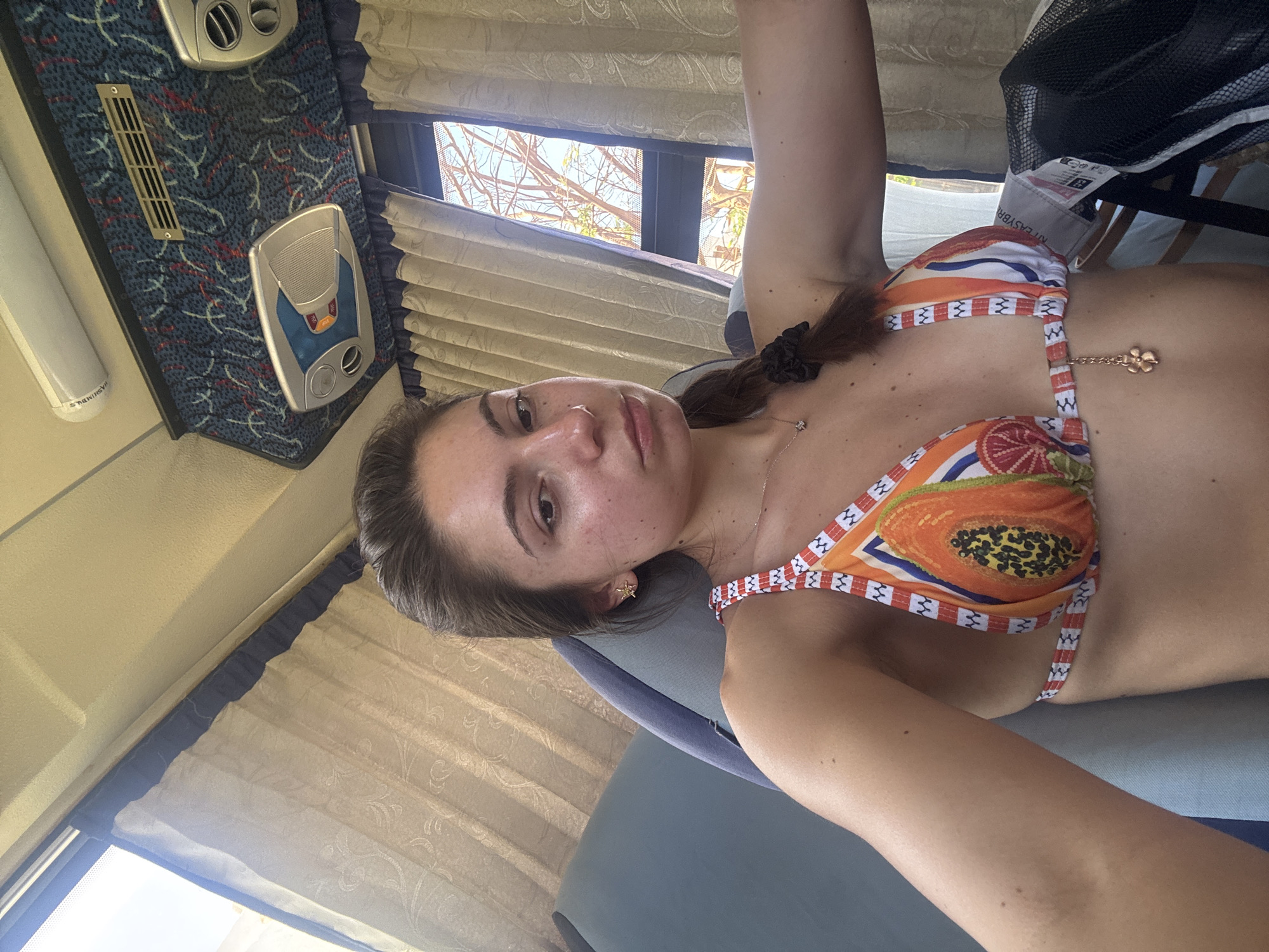 The image depicts a young woman wearing a bikini top with a papaya pattern, lying on a seat with a light blue or gray upholstery. The setting appears to be indoors, possibly on a bus or a recreational vehicle, as suggested by the patterned interior, window with curtains, and overhead control panel. The location is in the city of Unknown, Unknown. The woman is looking at the camera with a neutral expression. A window is visible in the background, and the natural lighting indicates it is daytime.