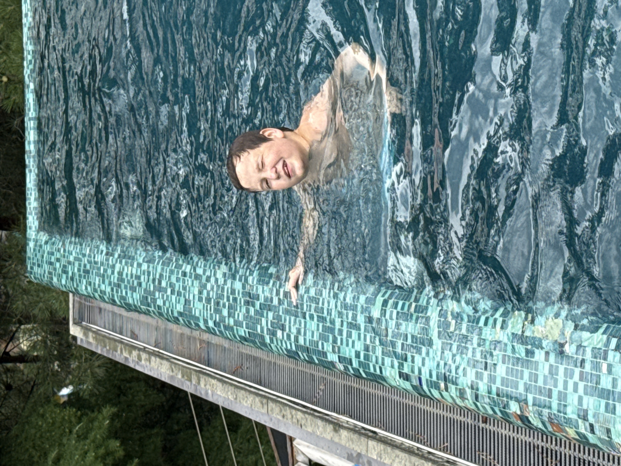 A young boy with brown hair is in a swimming pool. He is partially submerged in the water and smiling, with his eyes closed or squinting. One of his hands is resting on the edge of the pool, which is lined with blue and green mosaic tiles. The pool appears to be an infinity pool, with a metal grate visible along the edge. Lush green trees and foliage are visible in the background. The scene suggests a recreational setting, possibly a private residence or a hotel in Yalta, Ukraine.