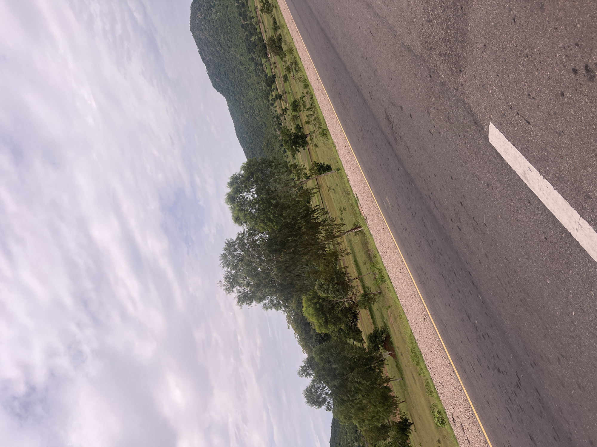 The image depicts a roadside scene in Minda, Nigeria, characterized by a paved road running alongside a green, hilly landscape dotted with trees. The perspective is from a low angle, looking diagonally across the road and into the landscape.
The road occupies the right side of the frame, its asphalt surface showing some wear and tear, with a white line marking the lane. A yellow line borders the road on the left. Beyond the road, a strip of gravel and then a grassy field with scattered trees stretches into the distance. In the background, a lush, green hill rises, covered in dense vegetation. The sky above is overcast with wispy clouds, suggesting a daytime scene, possibly in the late morning or afternoon.
The overall impression is one of a rural or semi-rural setting, with a sense of openness and natural beauty. There are no people or specific objects that indicate a particular event or activity. The environment appears tranquil, with no obvious signs of human activity beyond the road itself. The image captures the essence of the Nigerian landscape, with its vibrant greenery and rolling hills.
