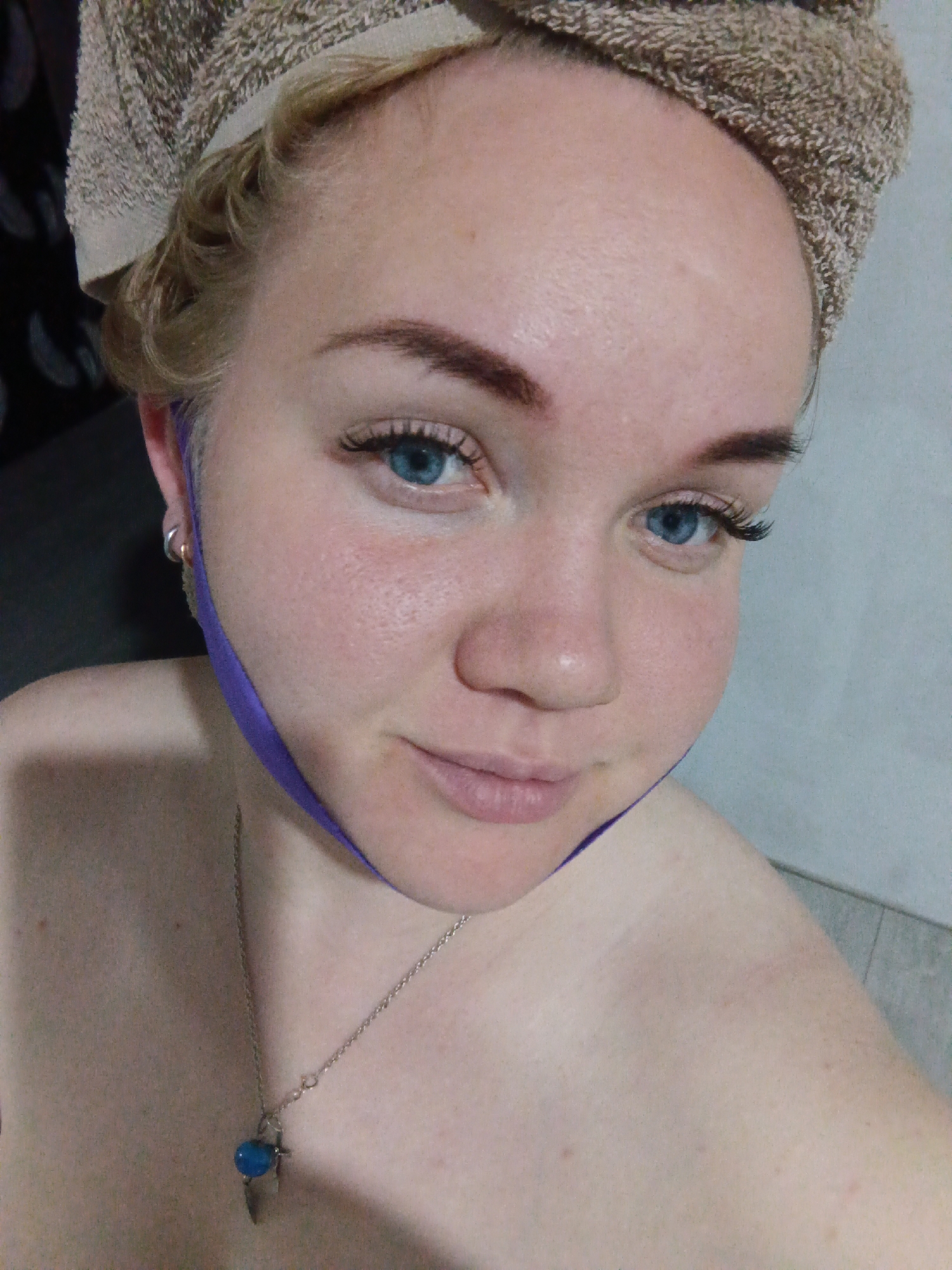 A fair-skinned woman with blonde hair wrapped in a towel is shown from the chest up. She has blue eyes, and a purple strap is positioned across her left cheek. A silver necklace with a blue pendant is visible around her neck. The background is a blend of dark and light gray surfaces, possibly a bathroom. The location is unknown.