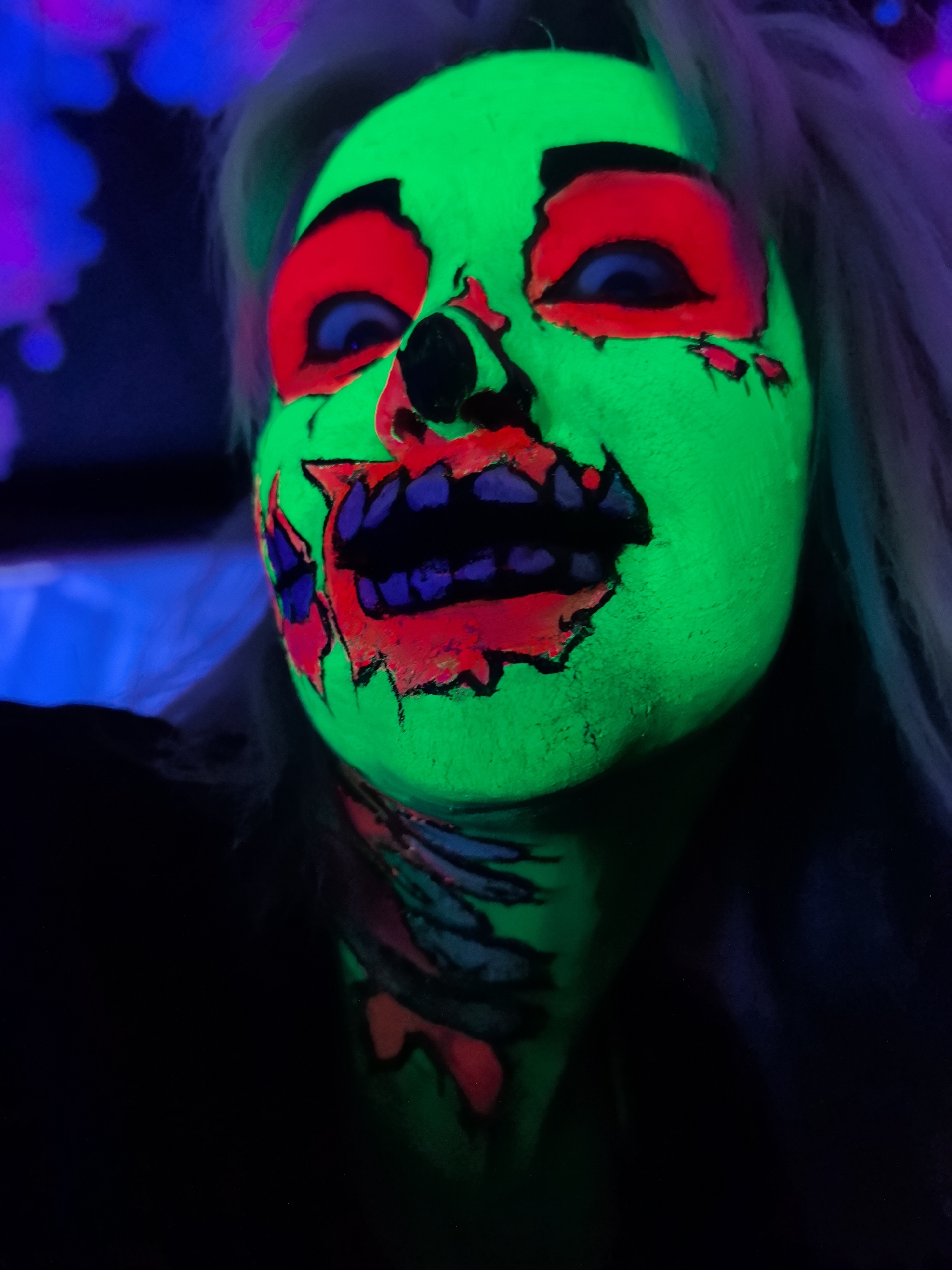 The image shows a close-up, head-and-shoulders portrait of a person with neon green face paint applied in a zombie or monster design. The face paint features bright red accents around the eyes and mouth, with black and purple details for the teeth and other facial features. The person's eyes are visible, appearing dark blue. Their hair is light-colored and extends to the sides. The background is dark and appears to have blurred purple and blue lights, suggesting a dimly lit environment, possibly a club or a party setting. There are no discernible features that would indicate the location in Noviy, Russia.