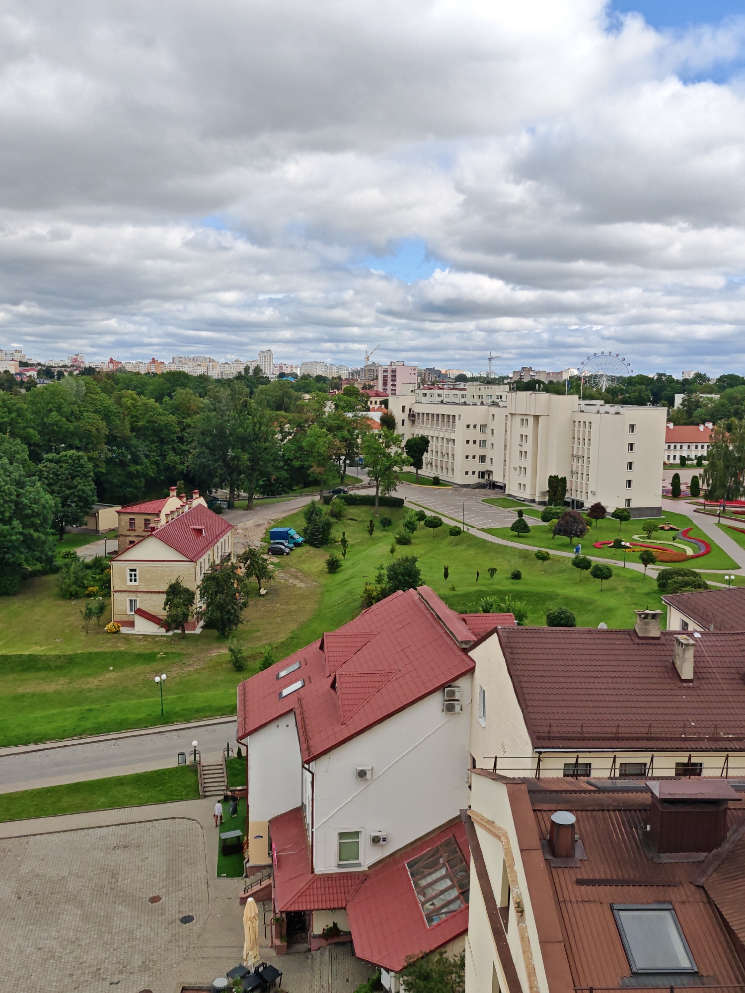 The image provides an aerial view of a cityscape in Hrodna, Belarus, featuring a complex of buildings, green spaces, and a Ferris wheel in the distance. In the foreground, a building with a distinctive red tiled roof dominates the lower section. This building has several windows, including skylights on its roof. Adjacent to it is another building, also with a red roof, painted in cream and white.
Moving towards the midground, a large, light-colored building with multiple stories and numerous windows stands prominently, surrounded by manicured green lawns and landscaping. Several trees are scattered throughout this area. A paved pathway leads to this building, and a colorful flowerbed forms a striking visual element. On the left side of the midground, a smaller building with a red roof is visible, with a road and parked vehicles in front of it.
The background reveals a more distant urban landscape with apartment buildings and infrastructure. A large Ferris wheel is a notable feature in the far background, suggesting a recreational area or amusement park. The sky is partially covered with clouds, indicating an overcast or partly cloudy day. The overall scene depicts an urban environment with a blend of modern architecture, green spaces, and recreational elements.