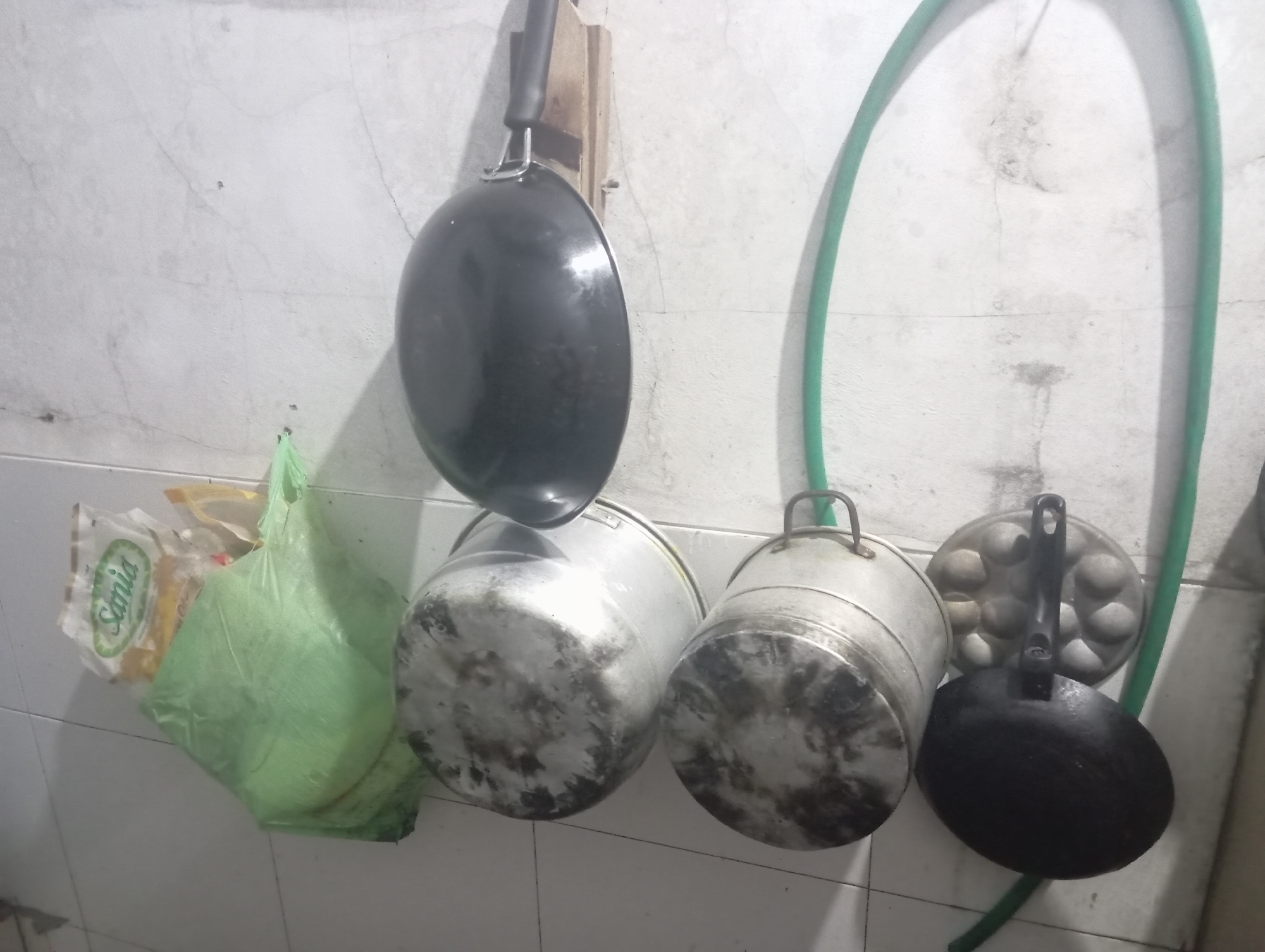 In Kutasari, Indonesia, a collection of kitchenware is displayed on a tiled wall. A black frying pan with a metal handle hangs from a wooden fixture. To its right, a green hose is coiled and hangs down. Next, a light green plastic bag containing packaged goods is suspended, with one package labeled "Sania" visible. Two large, metallic pots with handles are placed side-by-side, showing signs of use. Further to the right, a circular pan with indentations, likely for cooking small items like eggs, is mounted on the wall. Below this, another black frying pan is hanging. The overall scene depicts a utilitarian kitchen setting.