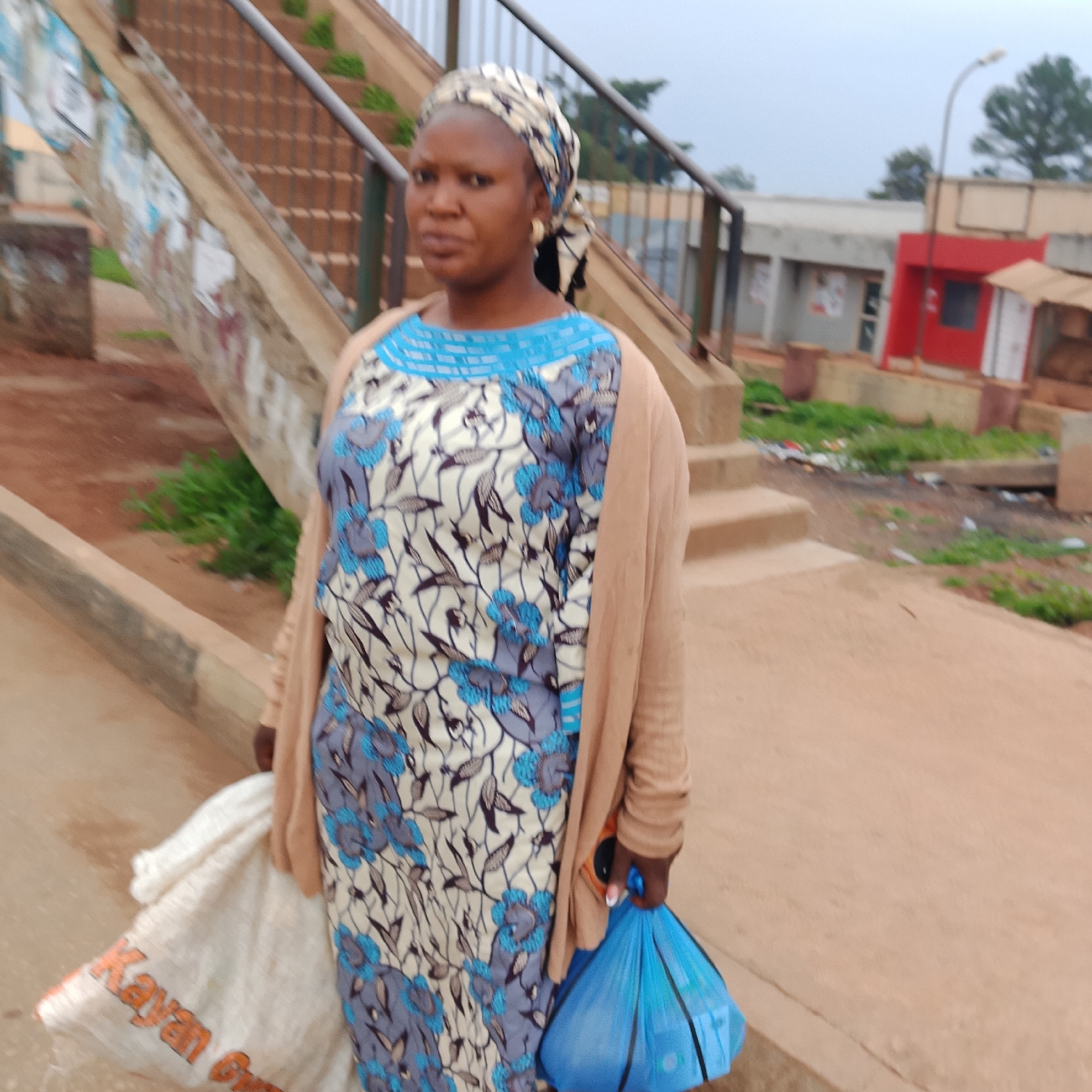In an outdoor, urban setting in Unknown, Unknown, a woman stands on a paved surface. She is wearing a patterned dress with blue and white floral designs and a beige cardigan. A patterned headscarf covers her hair. In her right hand, she holds a blue plastic bag. To her left, a large white cloth bag with orange lettering is visible, partially obscured by her body. Behind her, a set of stairs with a metal railing leads upwards. The background includes buildings with red and grey facades, a lamppost, and some greenery. The overall lighting suggests an overcast day.
