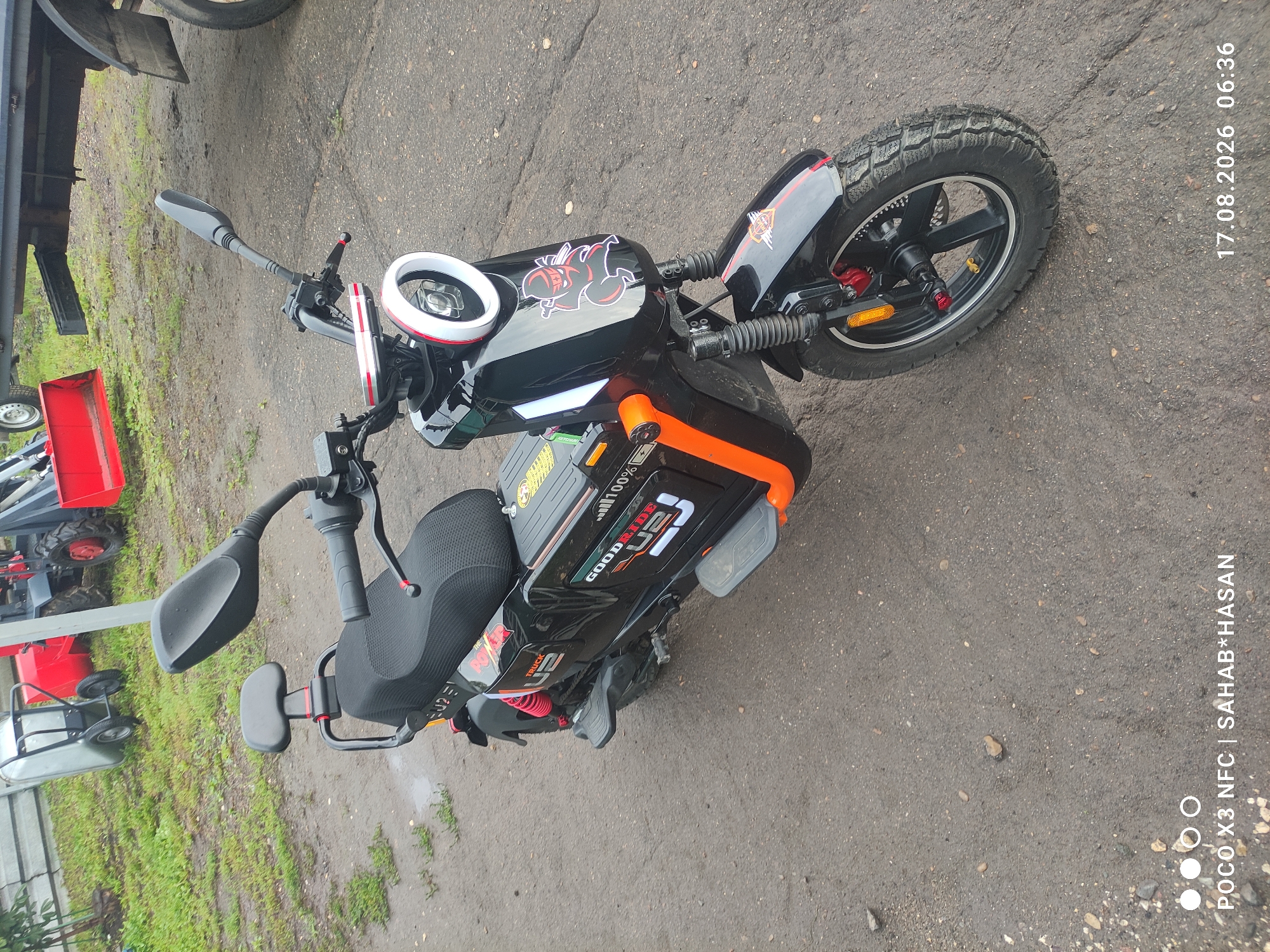 A black electric scooter with orange accents is parked on a wet, asphalt surface. The scooter has a round headlight with a white LED ring, a black seat with a small backrest, and "GOODRIDE U2" branding on its side. There is a sticker of a ninja on the front fairing. In the background, a wheelbarrow and a red tractor-like vehicle are visible on a grassy area with some mud. The scene appears to be outdoors, possibly in a rural or industrial setting. The image also contains a timestamp "17.08.2026 06:36" and a device indicator "POCO X3 NFC | SAHAB*HASAN". The presence of water and mud suggests recent rain. The specific location in Kostroma, Russia, cannot be determined from the image alone.