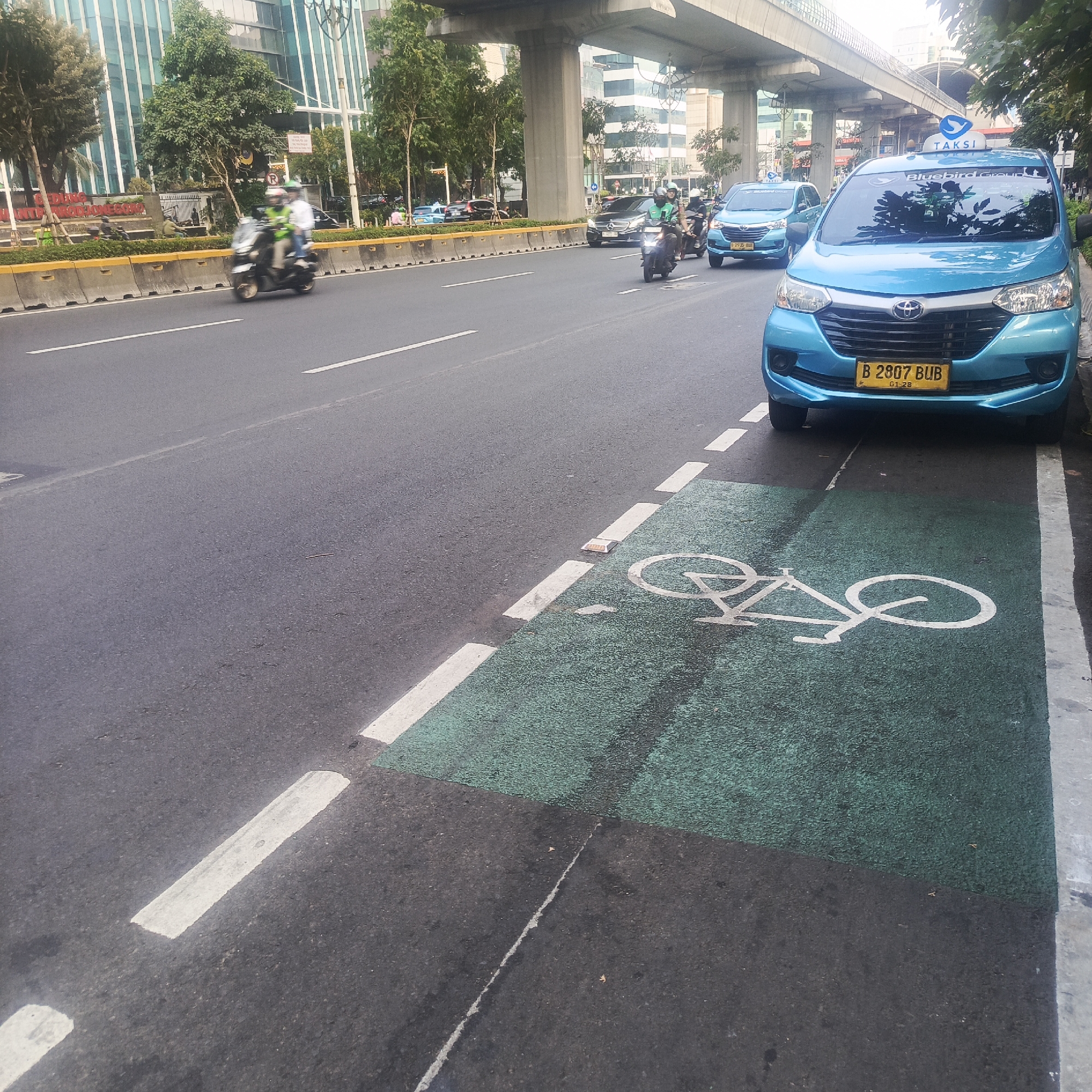 The image shows a street scene in South Jakarta, Indonesia, featuring a dedicated bicycle lane marked with green asphalt and a bicycle symbol. A blue Toyota Avanza taxi, with license plate B 2807 BUB, is parked partially within the bicycle lane, next to the curb. In the background, a highway overpass is visible, with traffic flowing on the road below. Two motorcyclists are also present, riding on the main road, one of whom is wearing a high-visibility green vest. The overall environment appears to be urban, with buildings and trees lining the street.