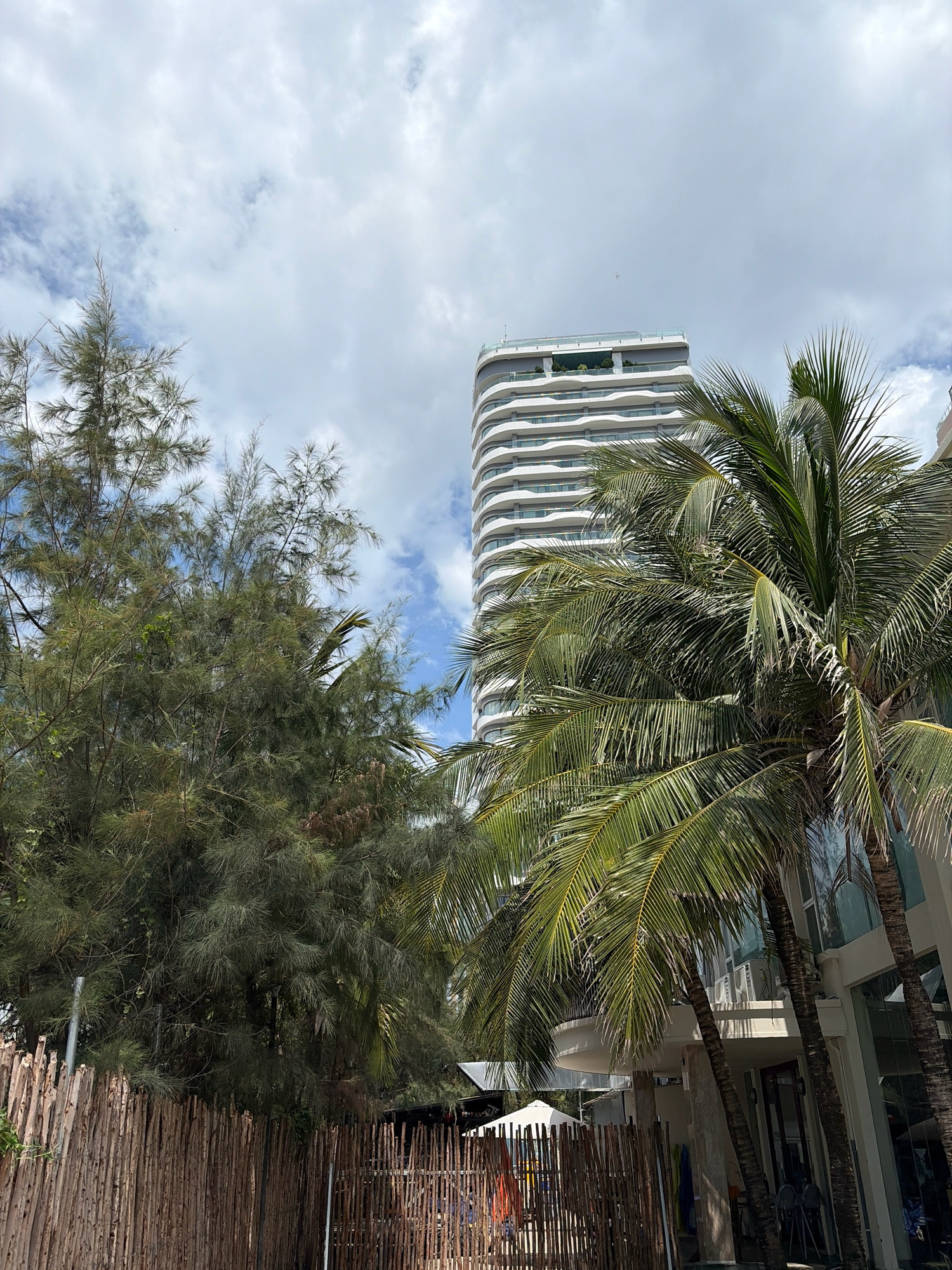 A tall, modern hotel with a white, wavy facade stands behind lush green palm trees and a grove of conifer trees. A rustic wooden fence runs along the foreground, with a white canopy tent visible behind it. The scene is set against a cloudy sky. This image was captured in the My Khe Beach area of Đà Nẵng, Vietnam.