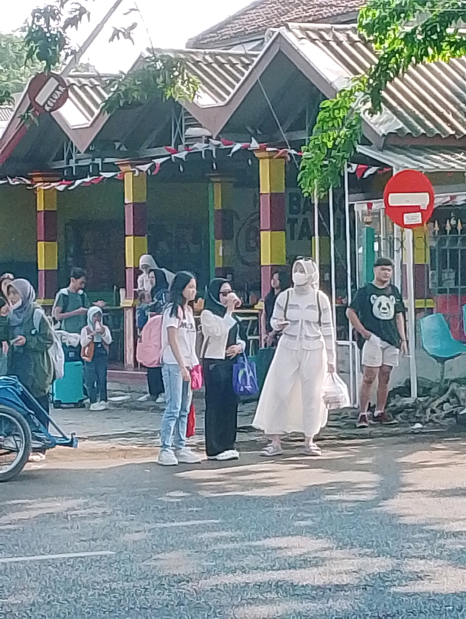 The image captures a scene on a street in Probolinggo, Indonesia, likely in front of a building with a distinctive roofline and colorful pillars. Several individuals are present. In the foreground, a person in a white outfit, possibly a woman, stands facing left, holding a plastic bag. To her right, a man wearing a dark t-shirt with a panda graphic and white shorts stands with his hand in his pocket, facing the viewer. To the left of the woman in white, a person with a pink backpack stands facing the viewer. Further to the left, another individual wearing a dark green shirt and a grey headscarf is visible. A child in a grey hoodie and dark pants is also present, holding a bottle. In the background, more people are visible, and a teal suitcase is on the ground. A red circular sign with a white horizontal bar is mounted on a pole. A blue bicycle rickshaw is parked on the left side of the frame. The environment appears to be outdoors, with some foliage visible, and the ground is paved. The scene suggests a gathering or waiting area.