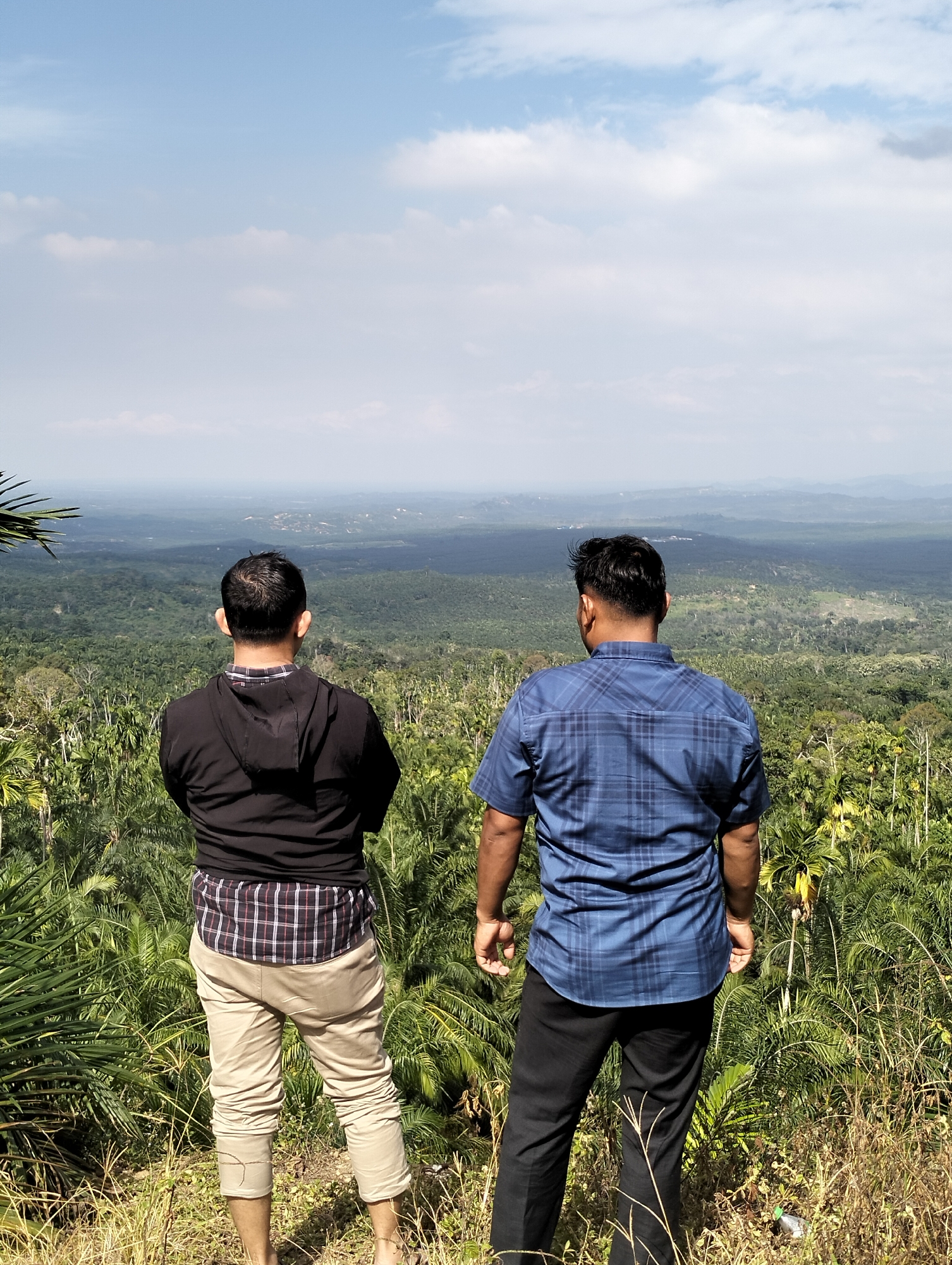 Two men stand with their backs to the camera, looking out over a lush, green landscape under a partly cloudy sky. The man on the left wears a black hooded jacket over a plaid shirt and light-colored pants. The man on the right is dressed in a blue plaid shirt and black pants. Both men appear to be in their late 20s or 30s. The foreground consists of dry grass and palm fronds. The background reveals a vast expanse of dense forest, with a hazy, mountainous horizon in the distance. The location is identified as Kilometer Delapan, Indonesia.