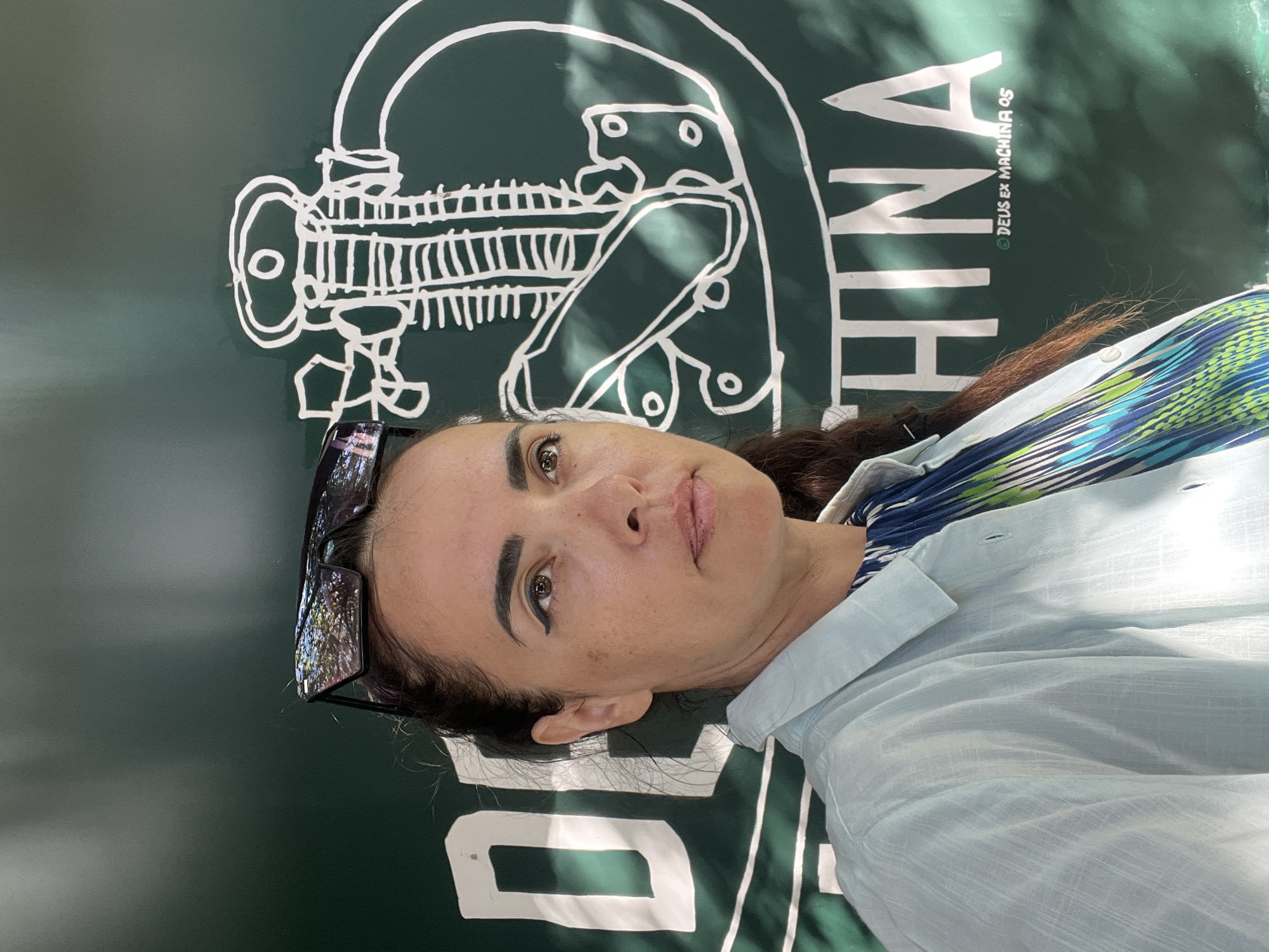 A woman stands in front of a green mural with white lettering and a drawing in Kuta Selatan, Indonesia. The woman has dark hair pulled back and is wearing sunglasses on her head. She is also wearing a light blue collared shirt over a patterned dress. The mural behind her appears to say "DEUS EX MACHINA" and features a stylized drawing of a mechanical object. Sunlight casts shadows across the mural and the woman's face.