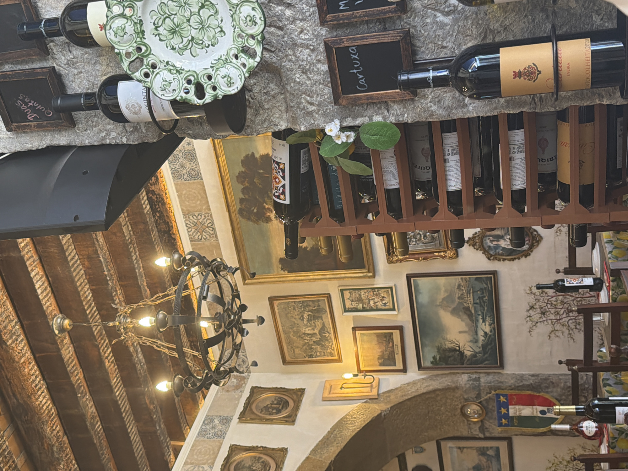 The image depicts the interior of a restaurant with a rustic and traditional ambiance, likely in Lisbon, Portugal, as suggested by the presence of Italian flag colors and the overall decor. The ceiling features exposed wooden beams and a wrought-iron chandelier. The walls are adorned with numerous framed paintings of varying sizes, some depicting landscapes and others featuring figures. A stone pillar on the right side of the image holds several bottles of wine, some displayed on a wooden wine rack and others standing on a shelf. Decorative plates, one with a green floral pattern, are also visible. The tables are set with tablecloths, and chairs are arranged around them. A flag with red, white, and green stripes, possibly the Italian flag, hangs on the wall near an arched doorway. The lighting is warm and inviting, contributing to the cozy atmosphere of the establishment.
