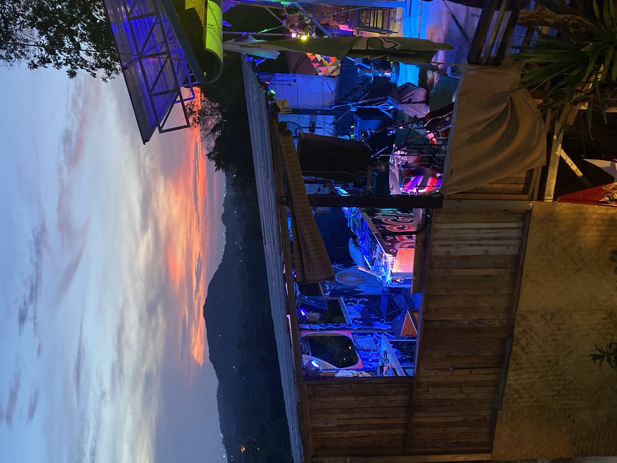 The image shows a brightly lit outdoor bar or performance space in Baan Don Sai, Thailand, at dusk. The bar has a corrugated metal roof and wooden construction. The space is decorated with colorful banners and lights. A mountain range with a vibrant orange and purple sunset sky is visible in the background. Musical instruments and equipment can be seen on a stage. Patrons are present.