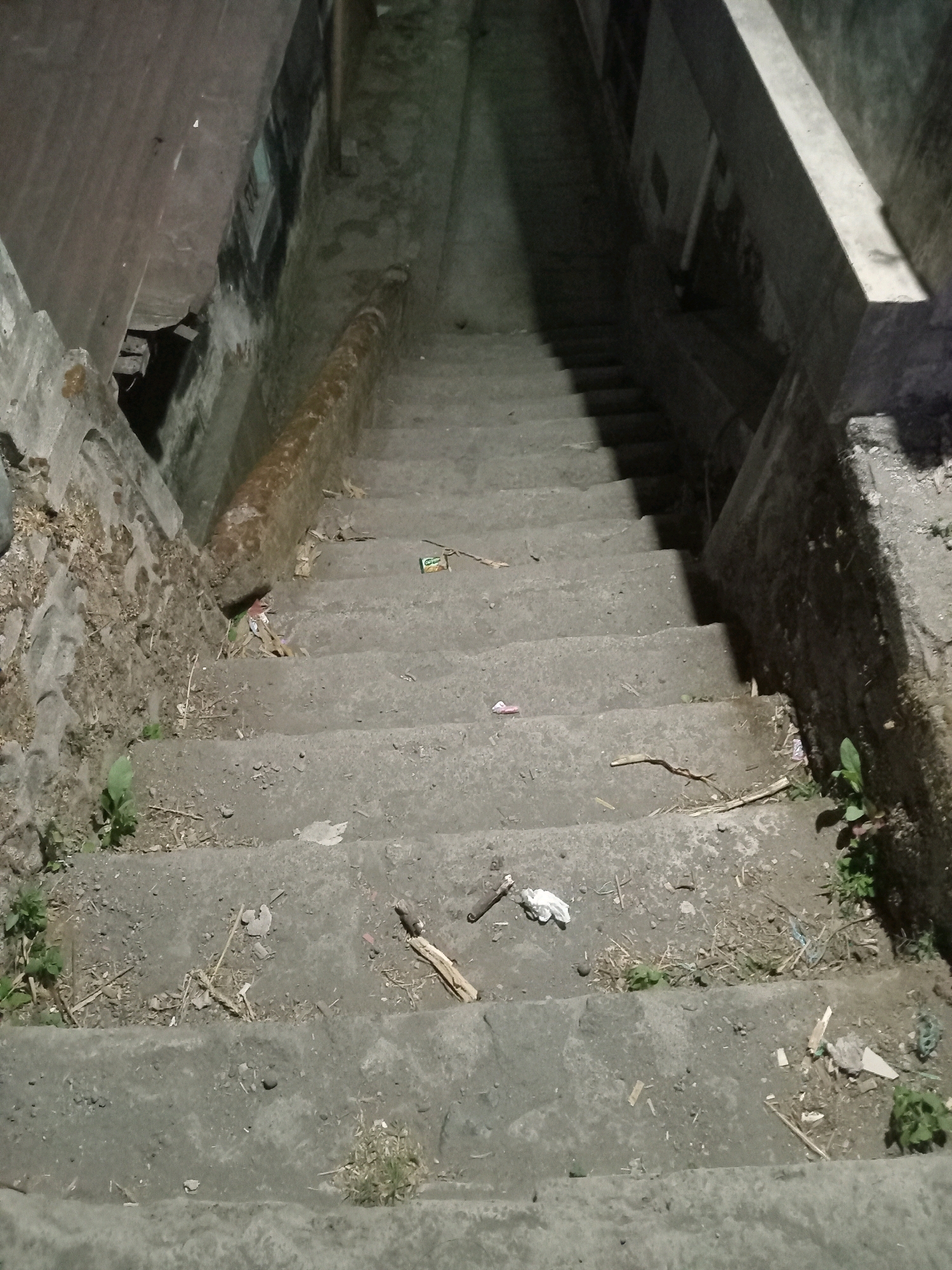 The image depicts a concrete staircase descending into darkness. The stairs are weathered and littered with debris, including dried leaves, twigs, and a few pieces of trash such as a crumpled wrapper and what appears to be a cigarette butt. Small patches of green vegetation are growing in the cracks and along the sides of the stairs. The lighting is dim, suggesting either nighttime or an enclosed, unlit space. The overall impression is one of neglect and disuse. The location is identified as Jurangjeru, Indonesia.