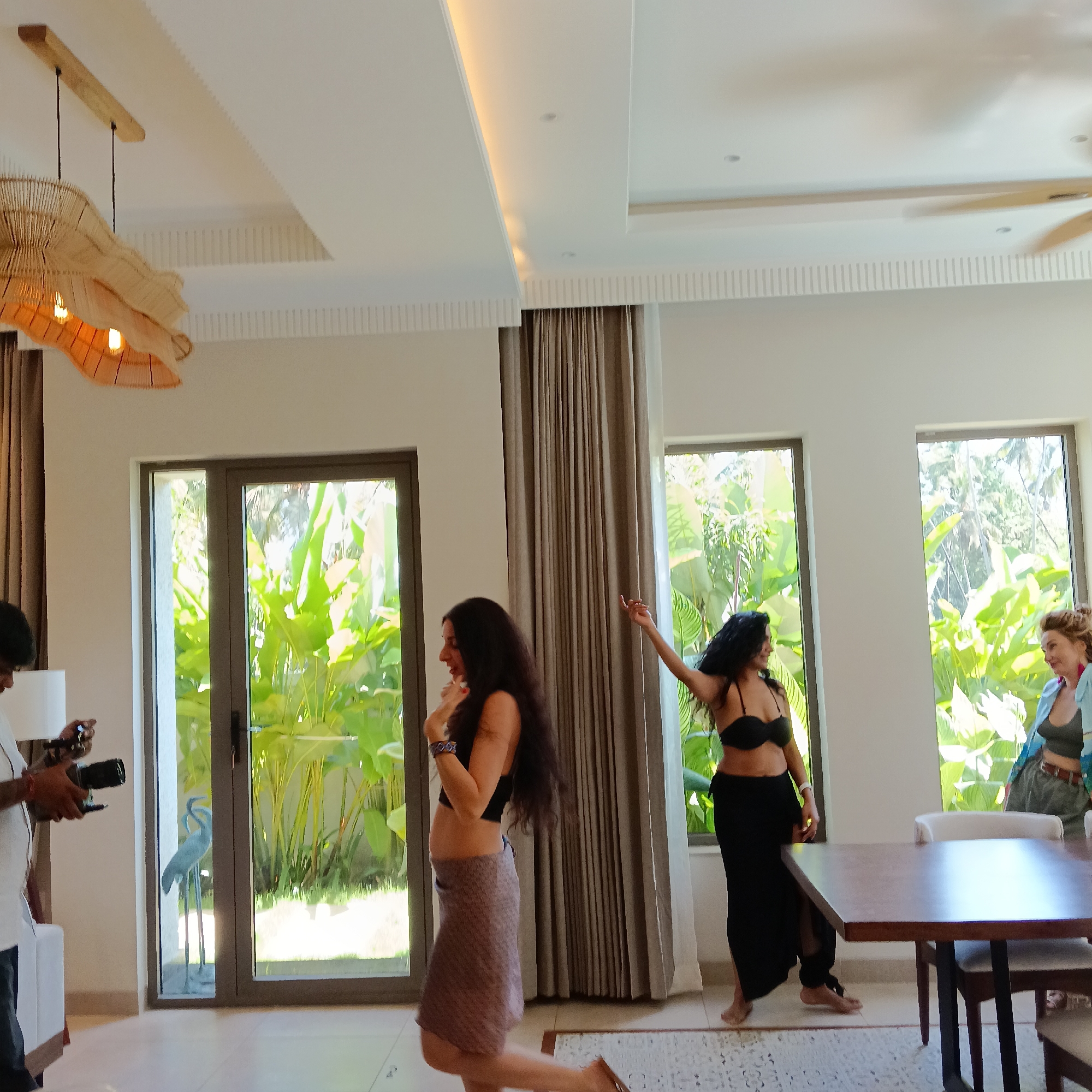 The scene appears to be a casual photoshoot taking place indoors, possibly within a villa or a large room in a residence. A male photographer is positioned on the left, holding a camera and focusing on a woman who is mid-pose. Another woman in a black bra top and long black skirt is striking a pose near a wooden table. The room is bright, with large windows showcasing lush green foliage outside, suggestive of a tropical environment. There are curtains partially drawn, and a unique woven light fixture hangs from the ceiling. The overall atmosphere suggests a relaxed, informal shoot. The location is given as Ashvem, India.