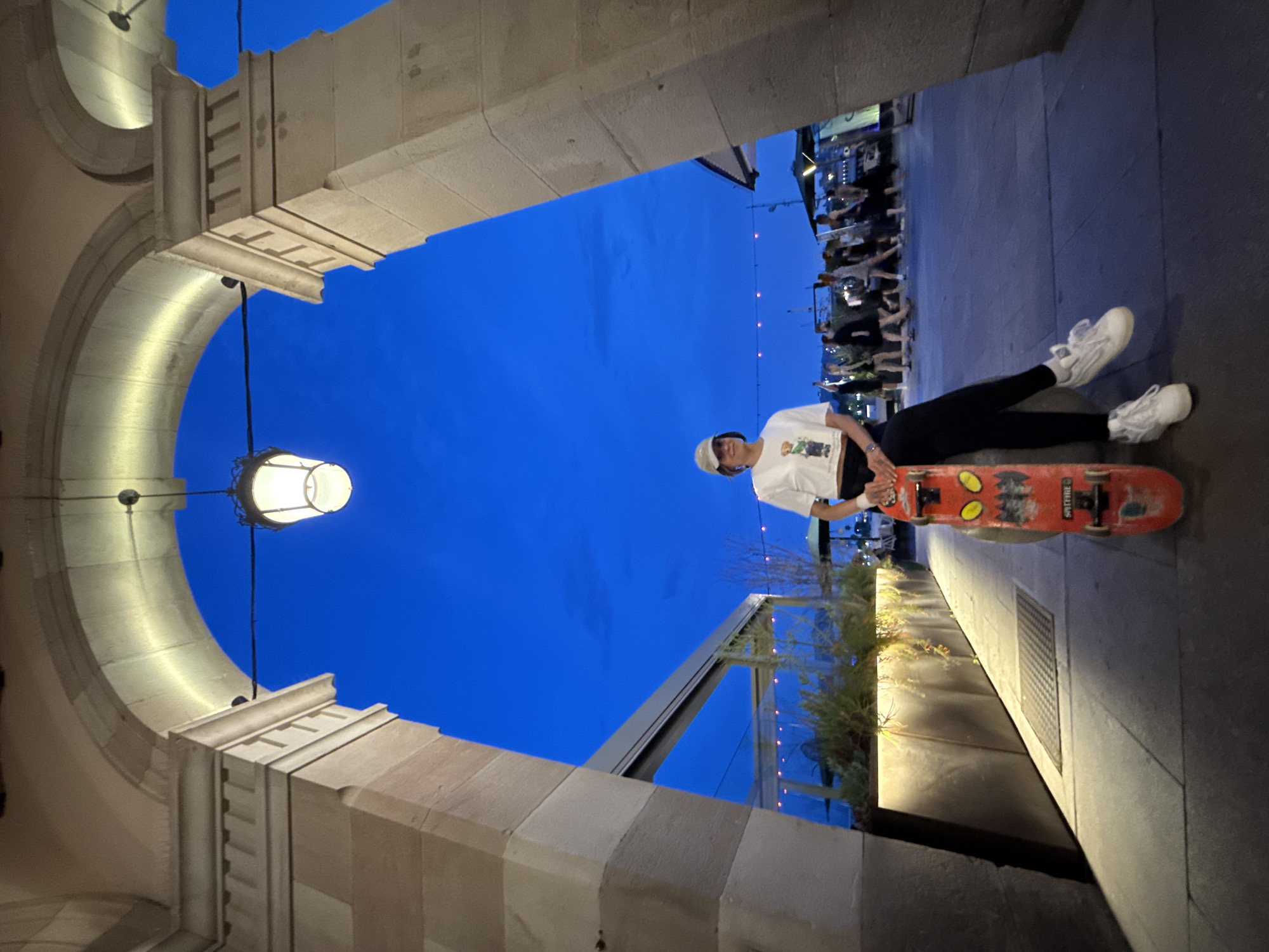 Here is a description of the image:
The photo is taken from a low angle, looking up through an arched opening of a building. The architecture suggests a location in a European city, possibly Barcelona, Spain, due to the building design. The archway is made of light-colored stone with decorative moldings and supports. A hanging light fixture is visible on the left side of the archway.
Beyond the arch, the sky is a deep blue, indicating that the photo was taken during twilight or early evening. To the right, a person is lying on a skateboard. The person is wearing white sneakers, black pants, and a white t-shirt. A few meters away, there are some plants. Further in the background, a group of people is observed, likely in a commercial establishment. A string of lights adds to the ambiance. The overall impression is that the photo was taken in an urban area, at dusk, where a person is leisurely resting on a skateboard while people around are enjoying the outdoor atmosphere.